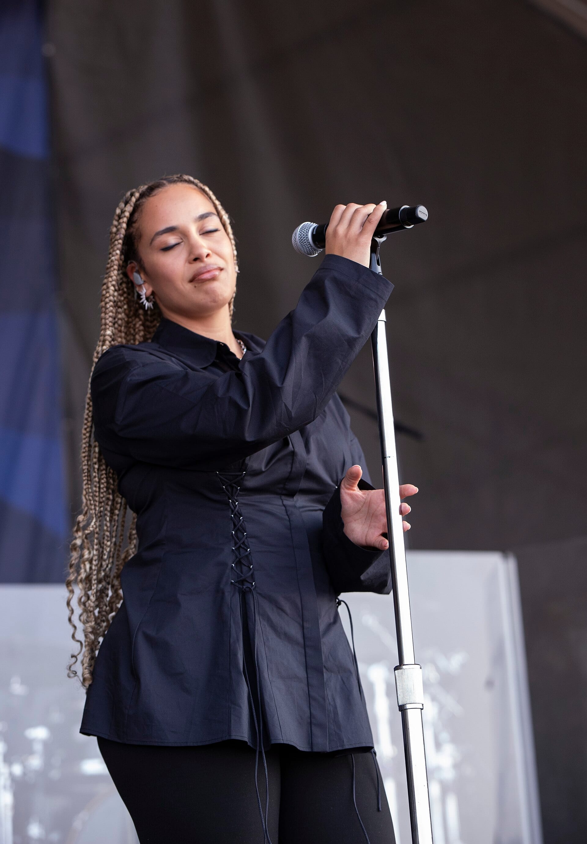 Jorja Smith sings with her eyes closed while holding a microphone stand, wearing a black corset-laced shirt with long braided hair falling past her shoulders.