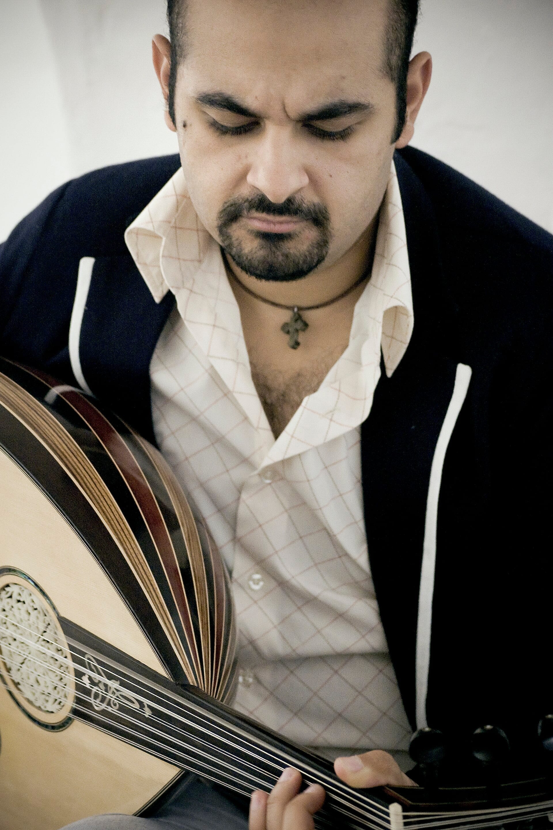 Joseph Tawadros looks down in concentration while playing the oud, wearing a dark blazer with white piping and a small cross pendant.