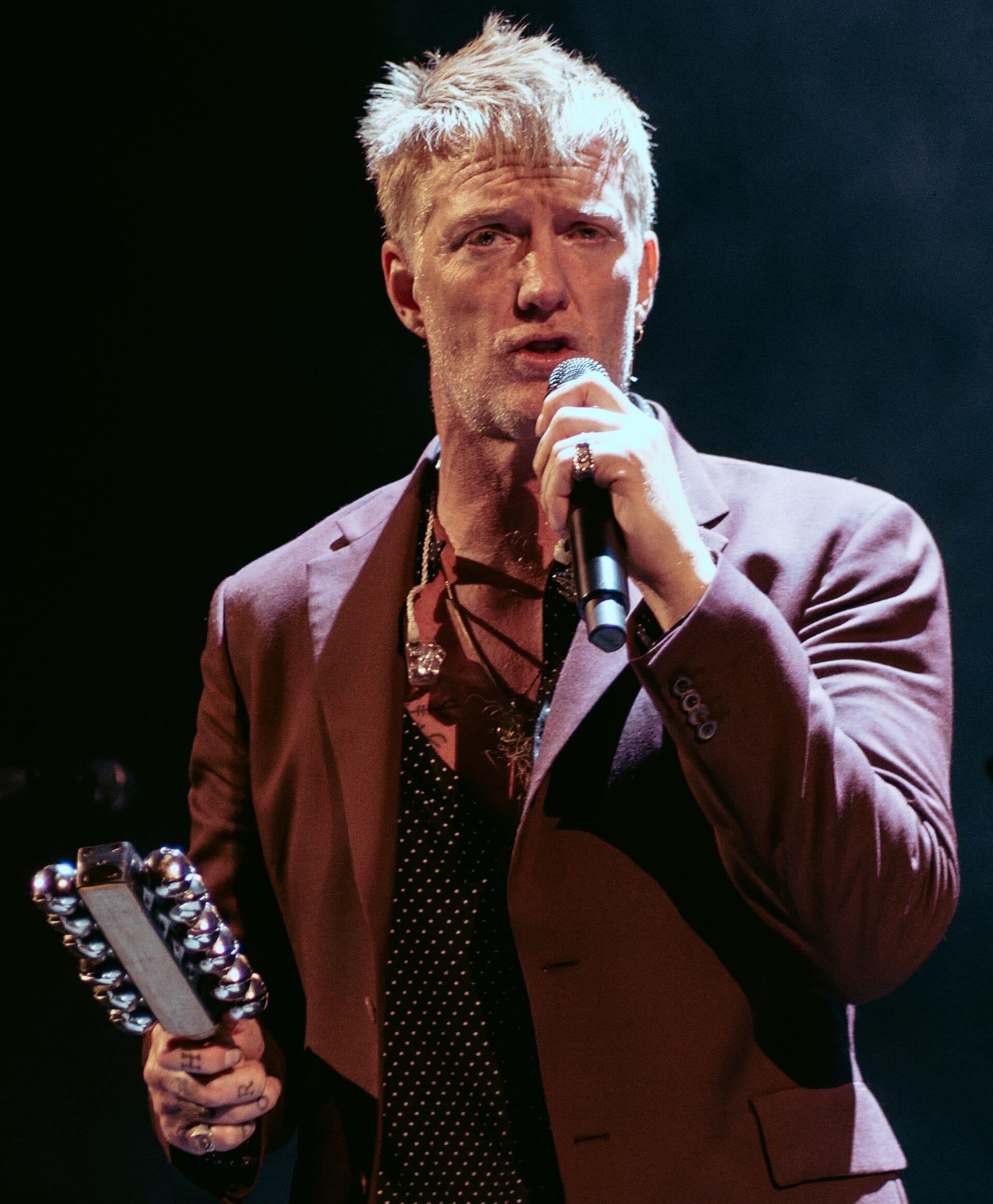 Josh Homme sings into a microphone with a tambourine in his free hand, in a burgundy jacket over a dark patterned shirt.