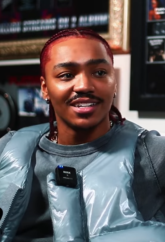 Josh Levi smiles mid-sentence in a radio studio, wearing a grey puffer jacket with a clip-on microphone, his braided hair dyed red.