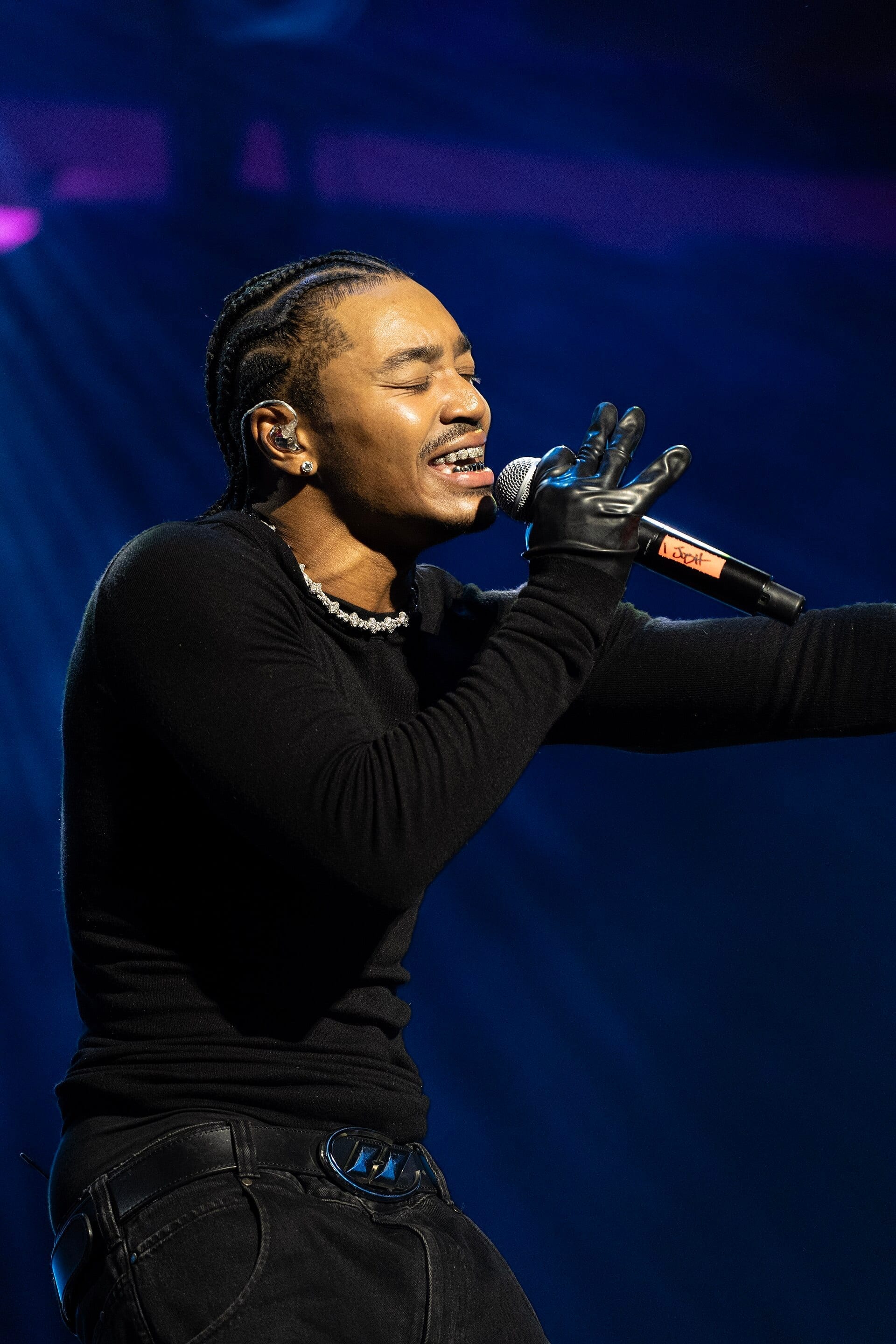 Josh Levi sings into a microphone with his eyes closed, wearing a diamond necklace and a black leather glove, lit by blue stage light.