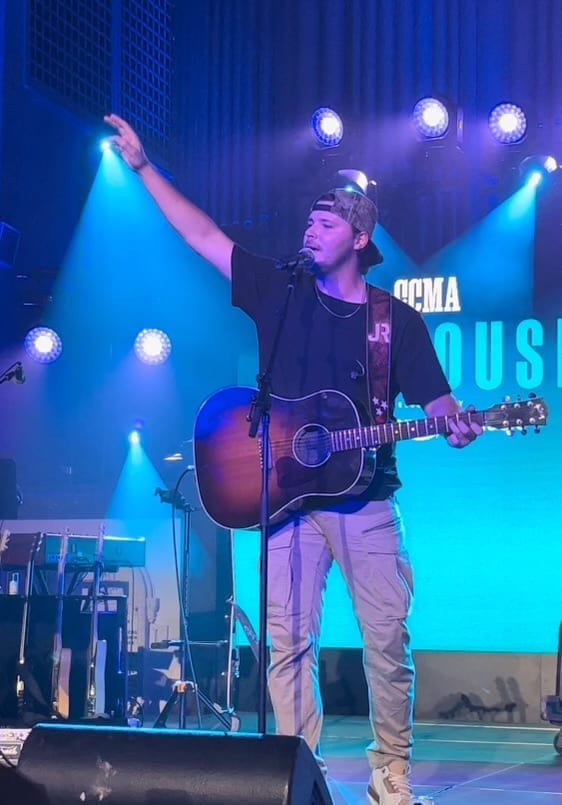 Josh Ross, wearing a backwards cap, raises one arm while singing into a microphone and holding an acoustic guitar on a blue-lit stage.