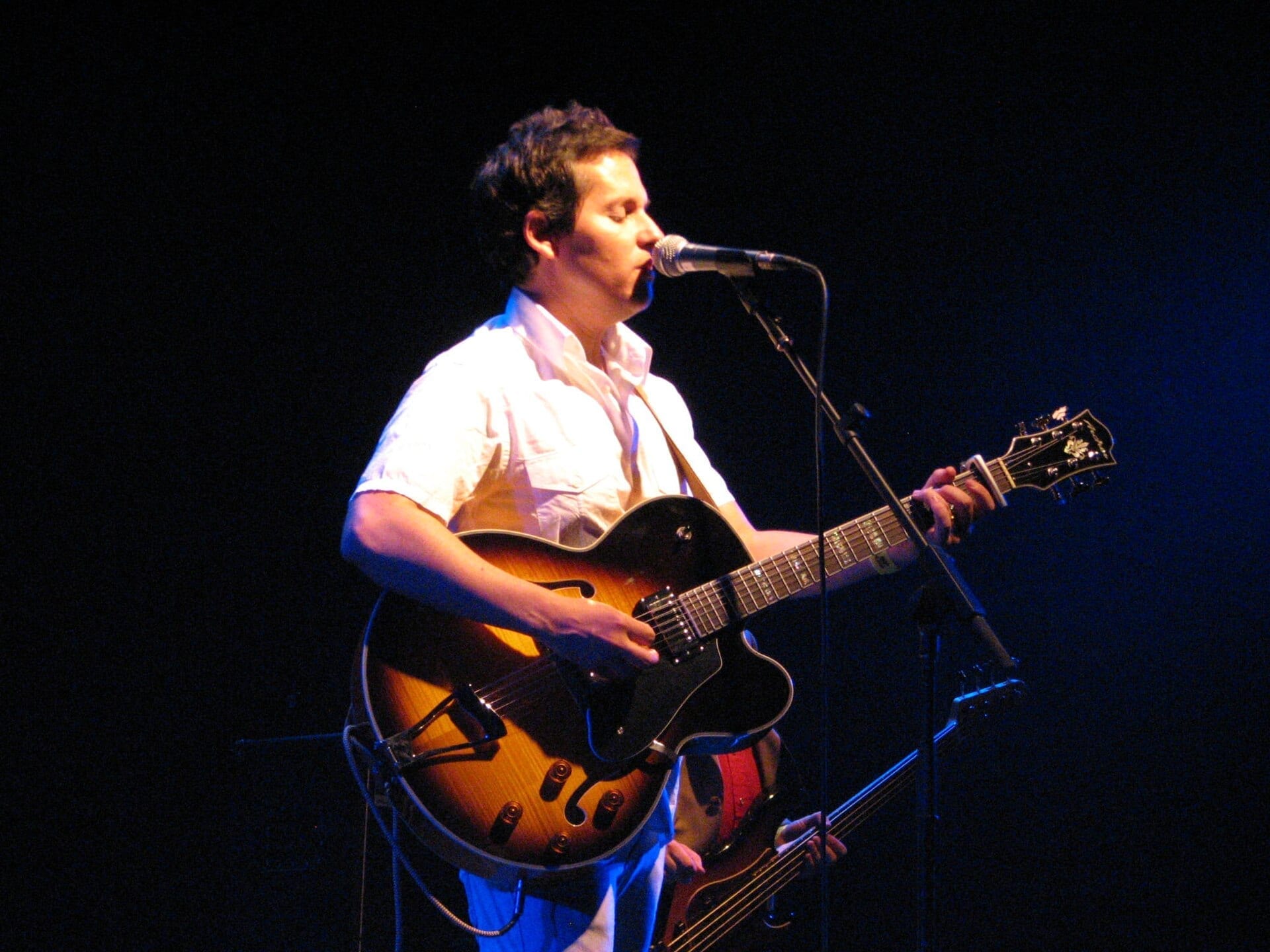 Josh Rouse singing into a microphone and playing an electric guitar on a dark stage.