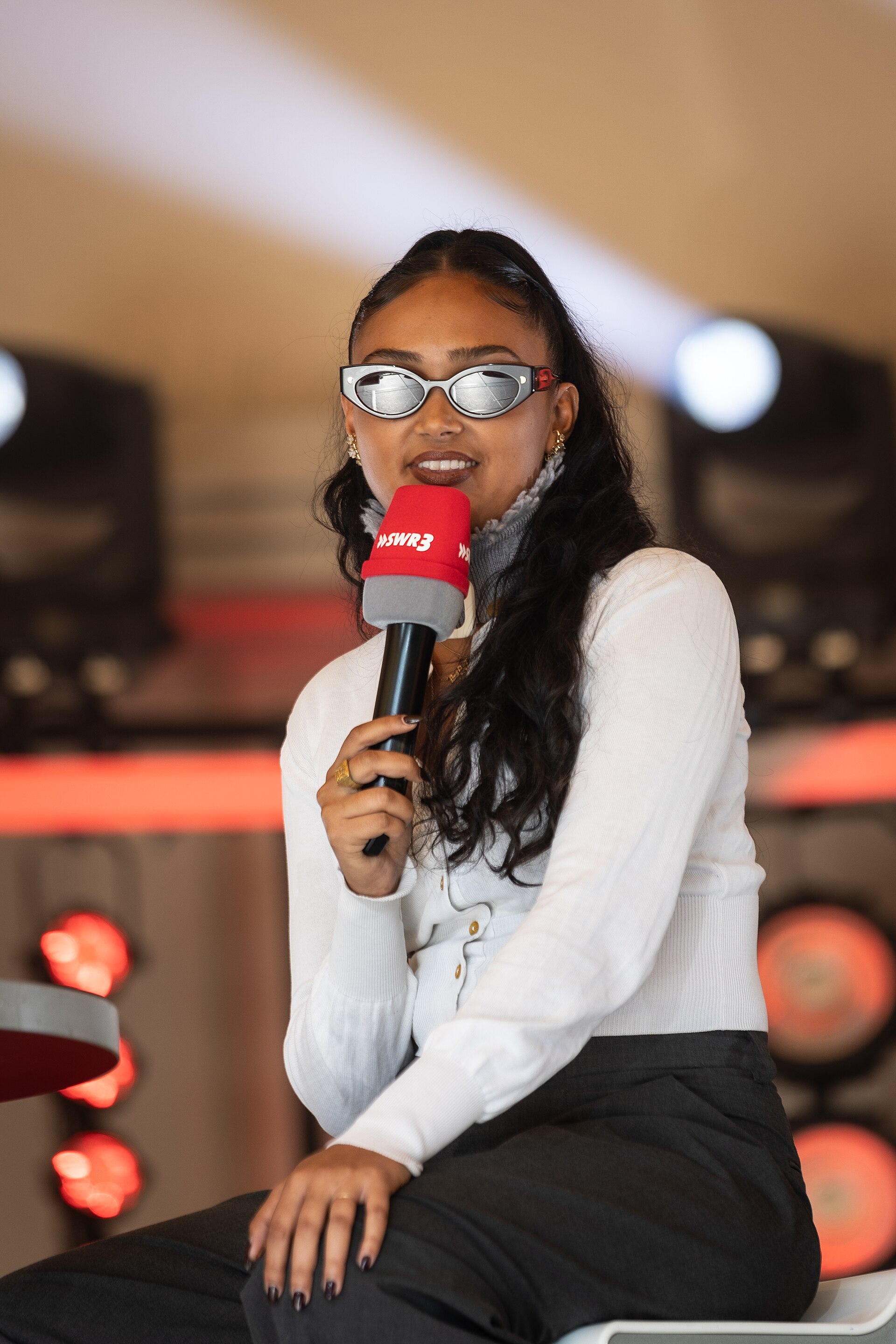 Joy Crookes sits with a red radio mic in one hand, in narrow silver sunglasses, a white cardigan and dark trousers.