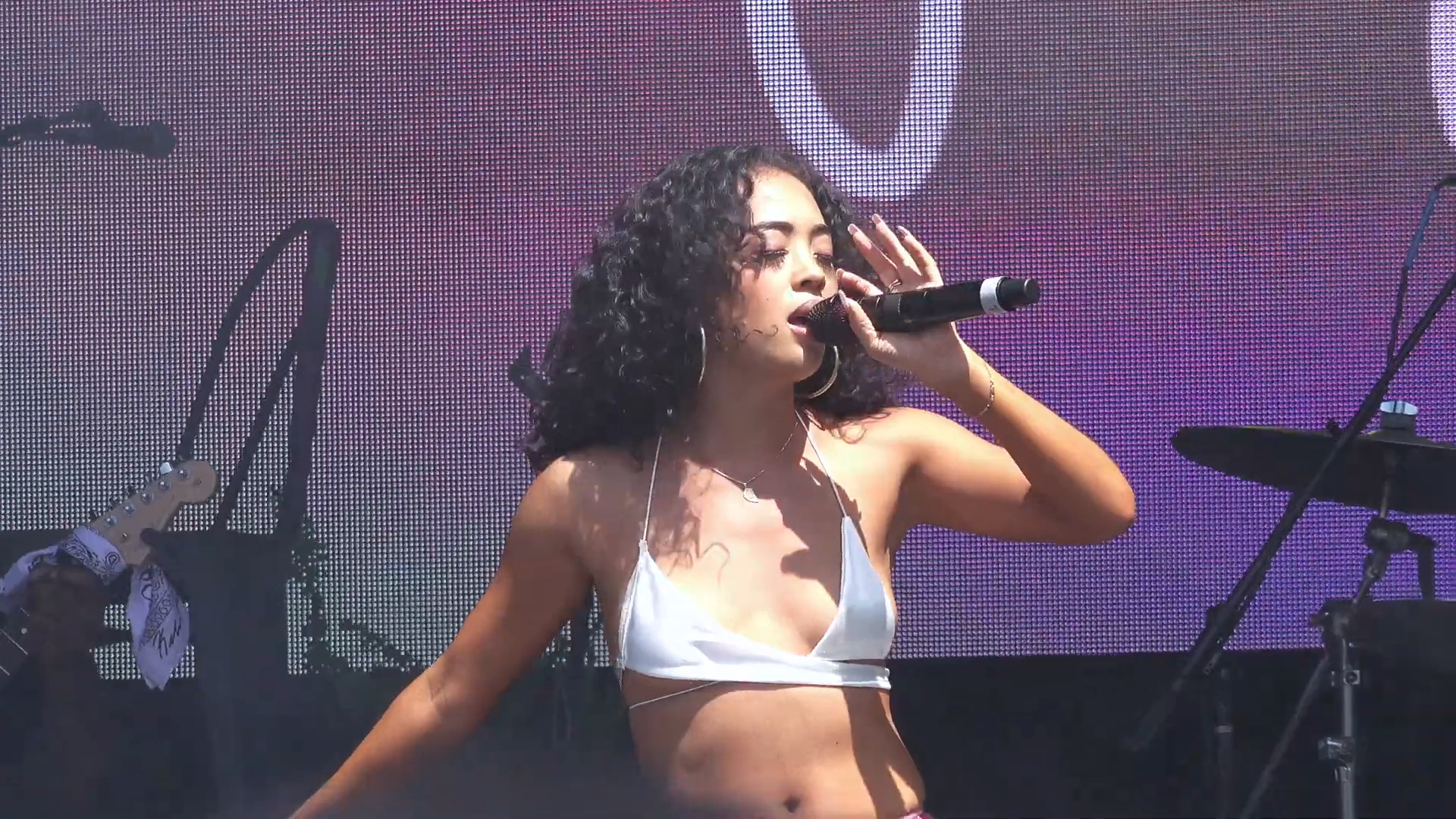 Joyce Wrice singing into a handheld microphone on an outdoor festival stage, curly hair loose, hoop earrings, a large video screen behind her