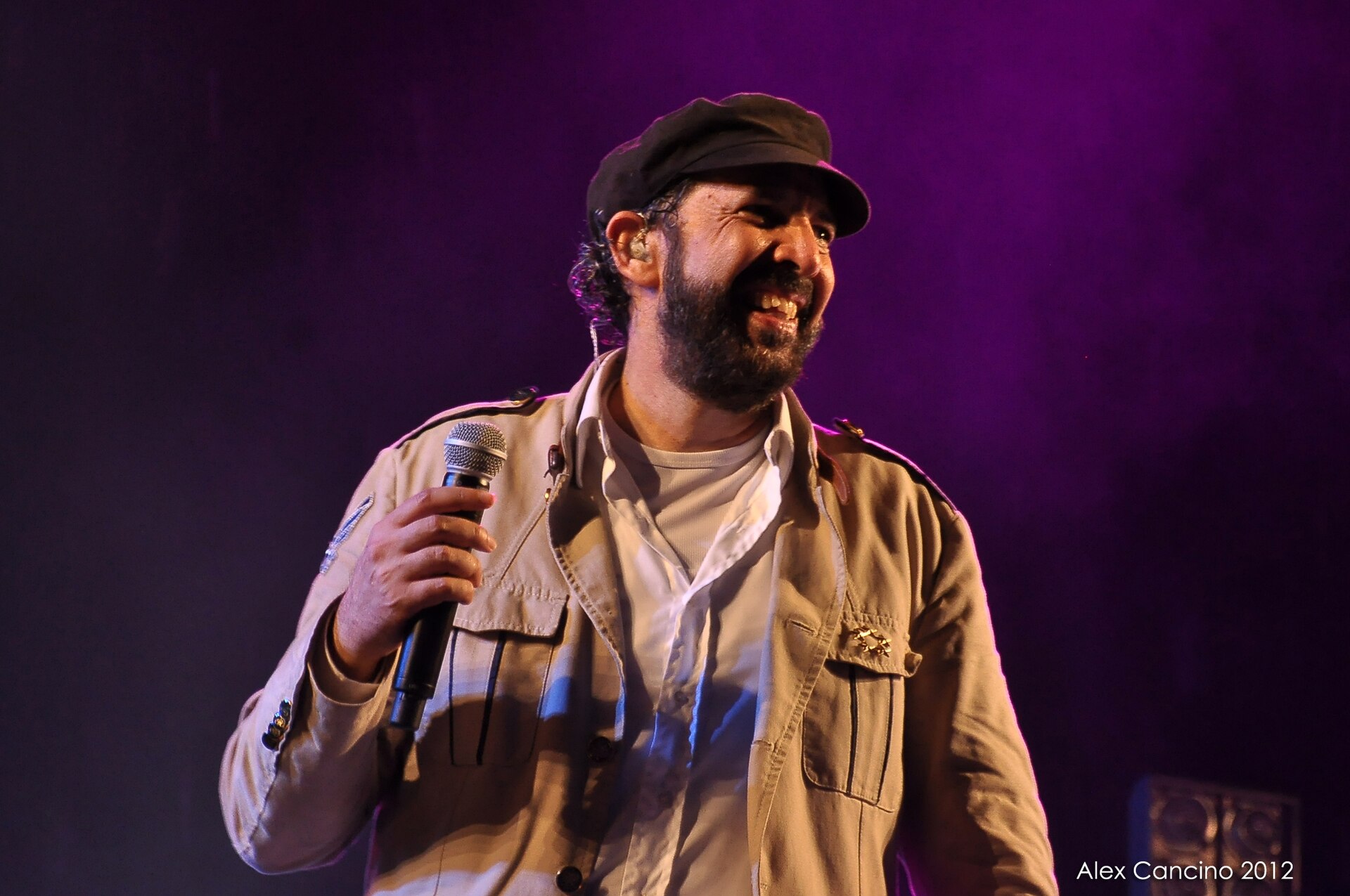 Juan Luis Guerra laughs on stage holding a microphone, in a flat cap and a pale utility jacket, lit purple from behind.