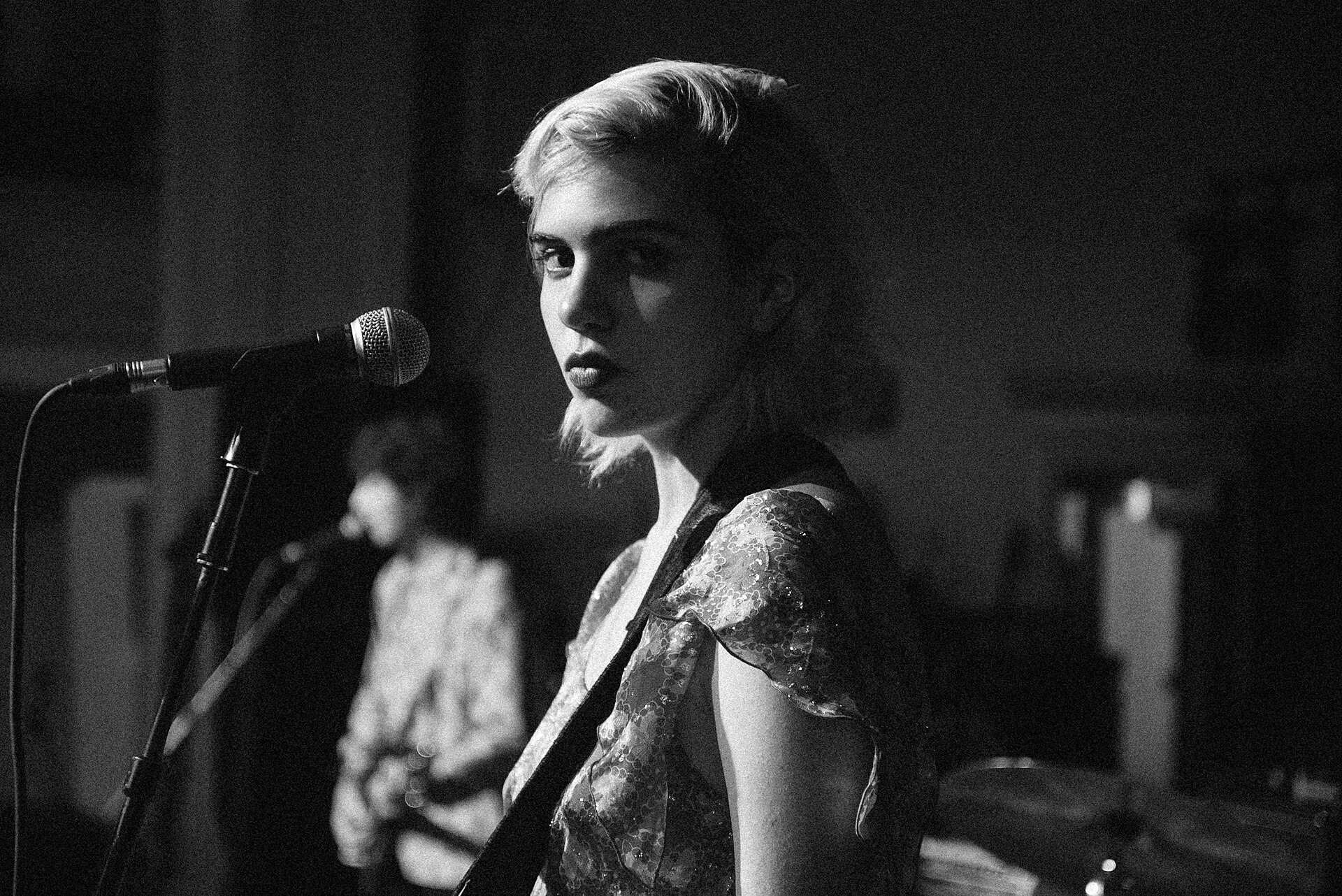 Julia Cumming stands at a microphone in black and white, bleached hair swept back, a guitar strap over a sheer patterned top, a bandmate out of focus behind her.