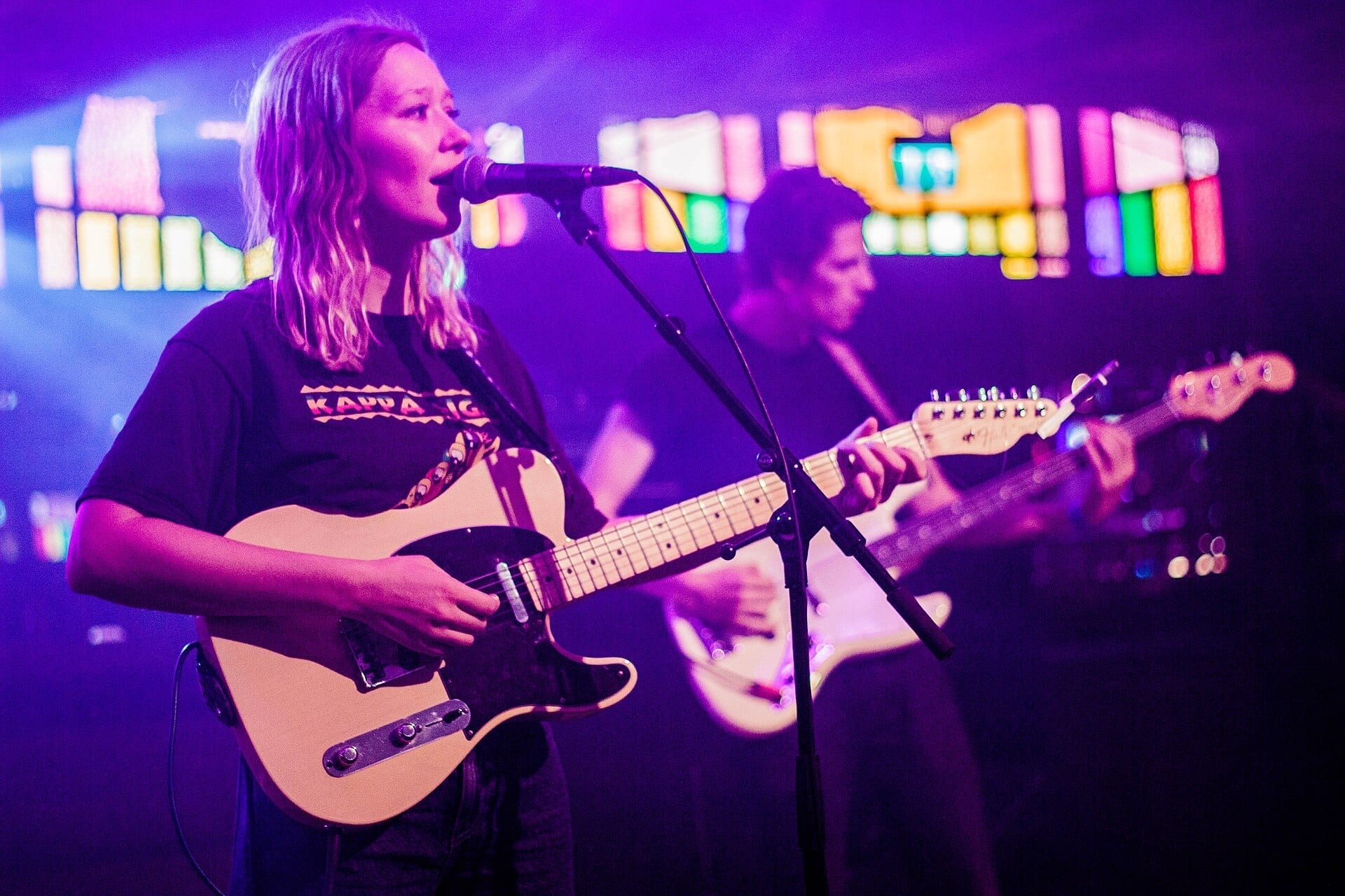Julia Jacklin sings into a microphone while playing an electric guitar on a colorfully lit stage, with a bandmate playing guitar behind her.
