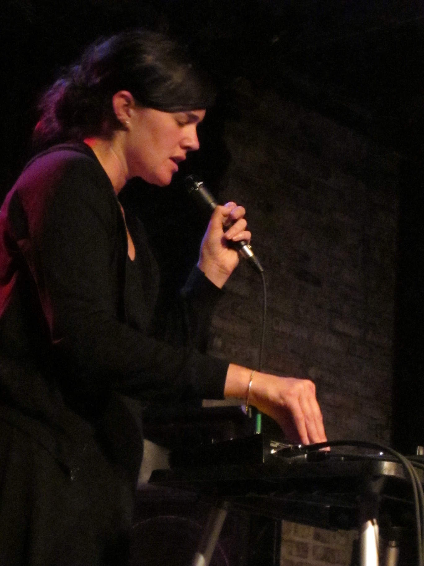 Julianna Barwick sings into a handheld microphone with her eyes down, her other hand resting on a pedal board, against a dark brick wall.