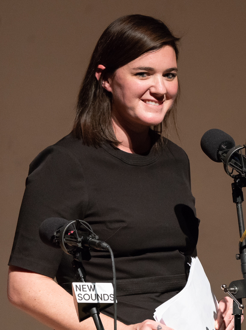 Julianna Barwick stands behind two microphones in a black top, holding sheets of paper, smiling off to one side.