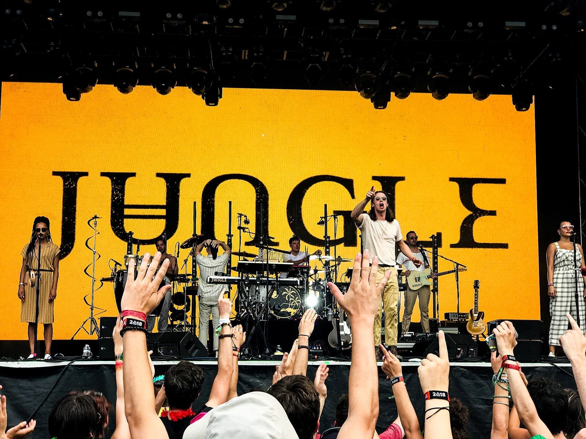 The band Jungle perform on a festival stage in front of a giant illuminated JUNGLE backdrop, with fans raising their hands in the crowd.
