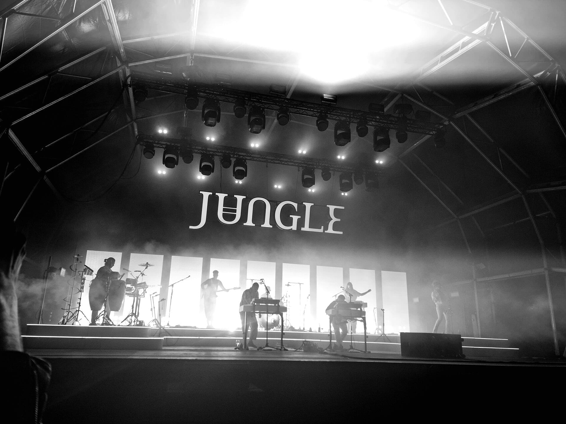 Black and white photo of Jungle's players in silhouette across a wide, hazy stage beneath an illuminated JUNGLE sign.