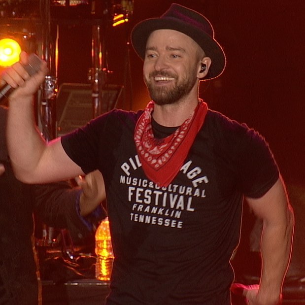 Justin Timberlake grins on a red-lit stage with a microphone raised in one hand, wearing a black festival T-shirt, red bandana and dark trilby.