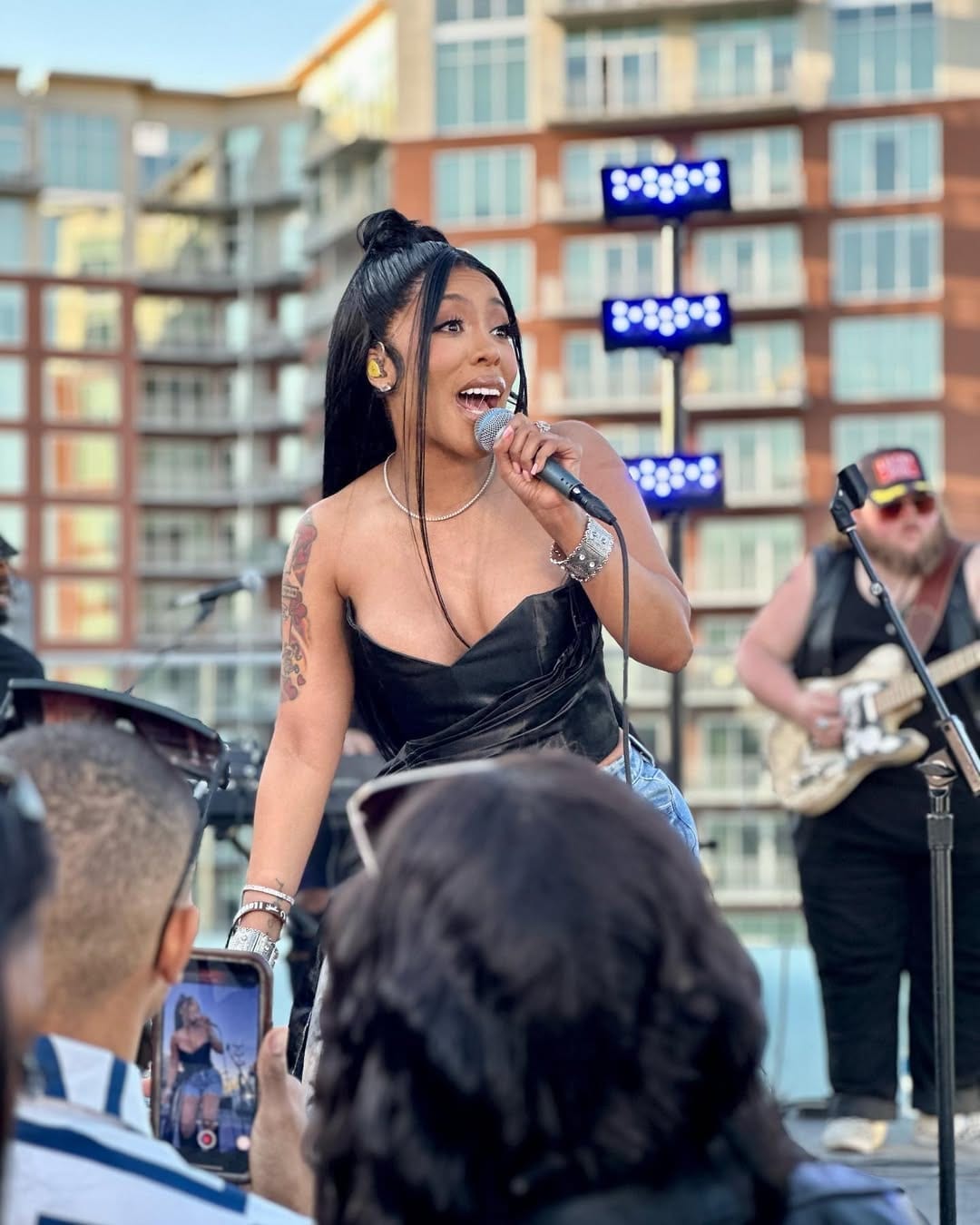 K. Michelle sings into a microphone during an outdoor performance, with a guitarist behind her.