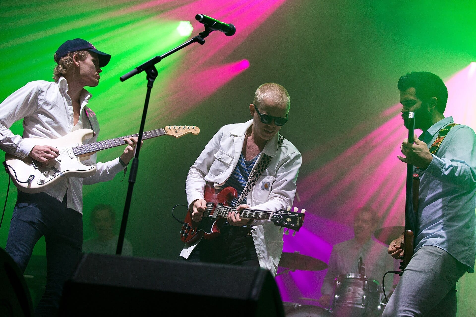 Three members of Kakkmaddafakka perform on stage under green and pink lights, two playing guitars around a microphone stand and a third playing bass at the side.