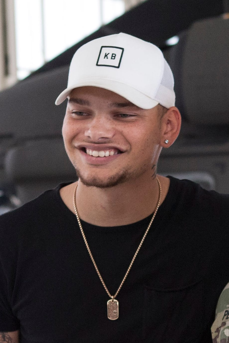 Kane Brown, wearing a white cap with his KB logo and a gold dog-tag necklace over a black t-shirt, smiles and looks off to the side.