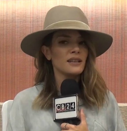 Kany Garcia speaks into a CB24 news microphone, wearing a wide-brimmed pale hat and a light shirt.