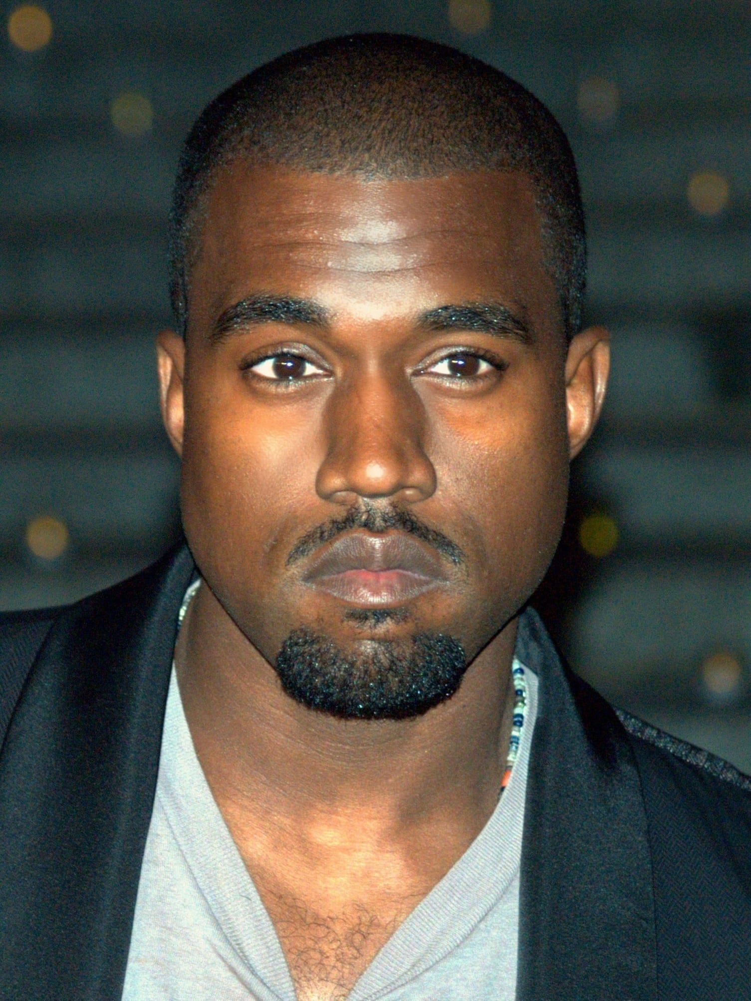 A close-up portrait of Kanye West with a short beard, looking directly at the camera in a dark jacket.