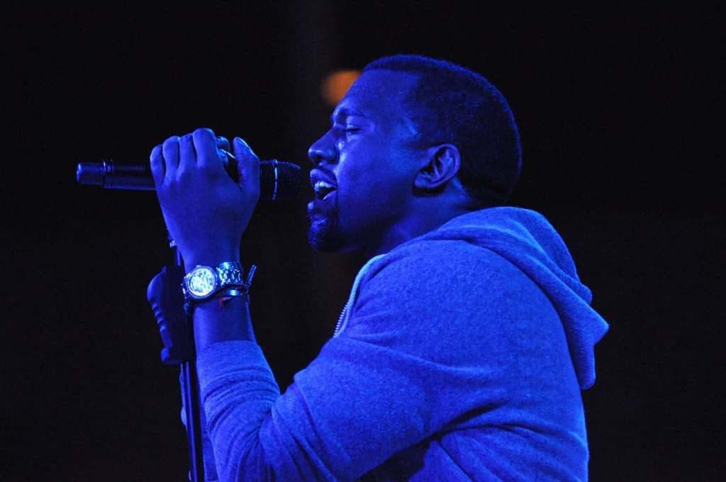 Kanye West in profile at a microphone stand, jaw set, lit from the side against a dark room.