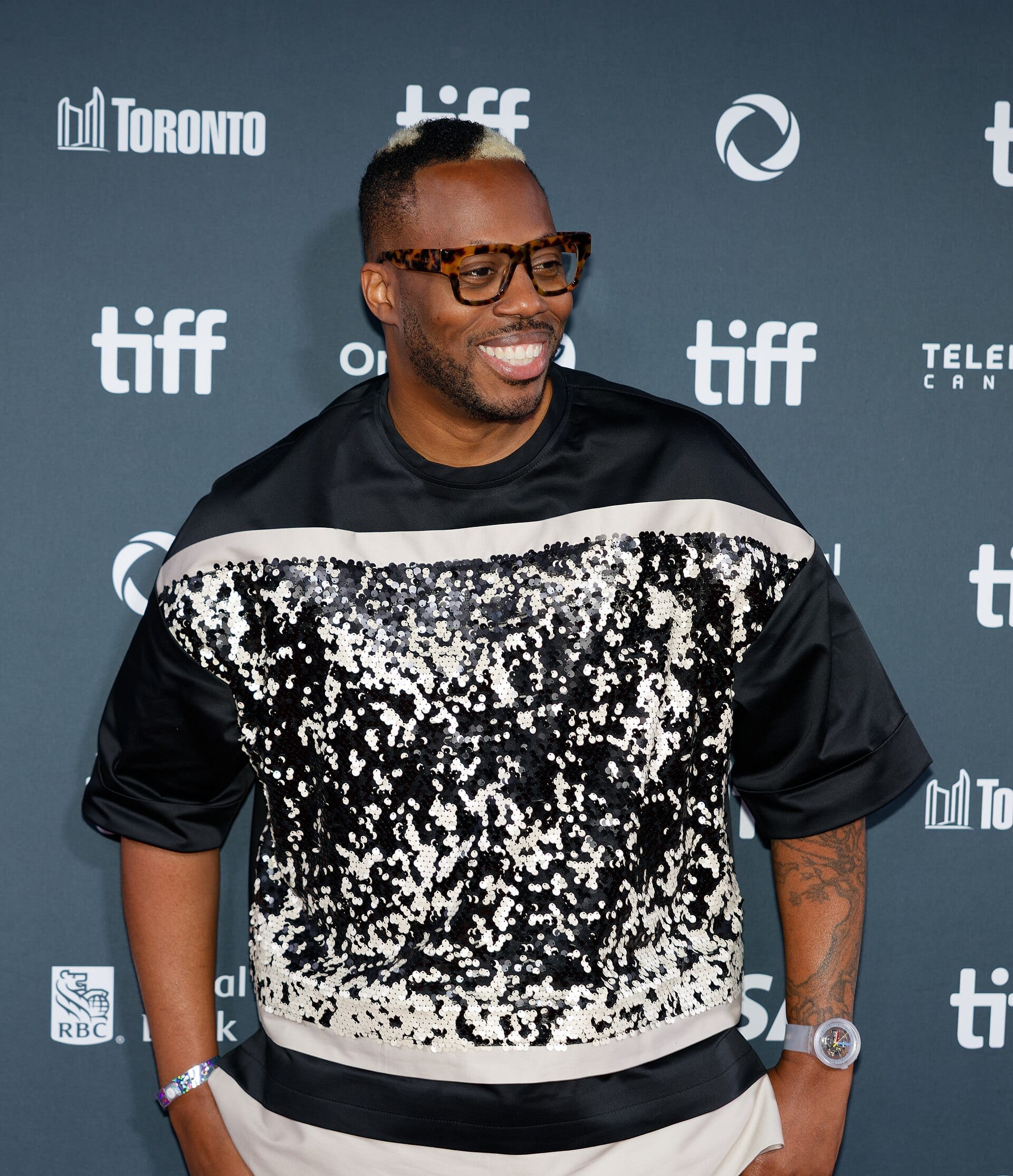 Kardinal Offishall grins on a festival step-and-repeat in tortoiseshell glasses and a black and cream sequinned top, his hair bleached blond at the front.