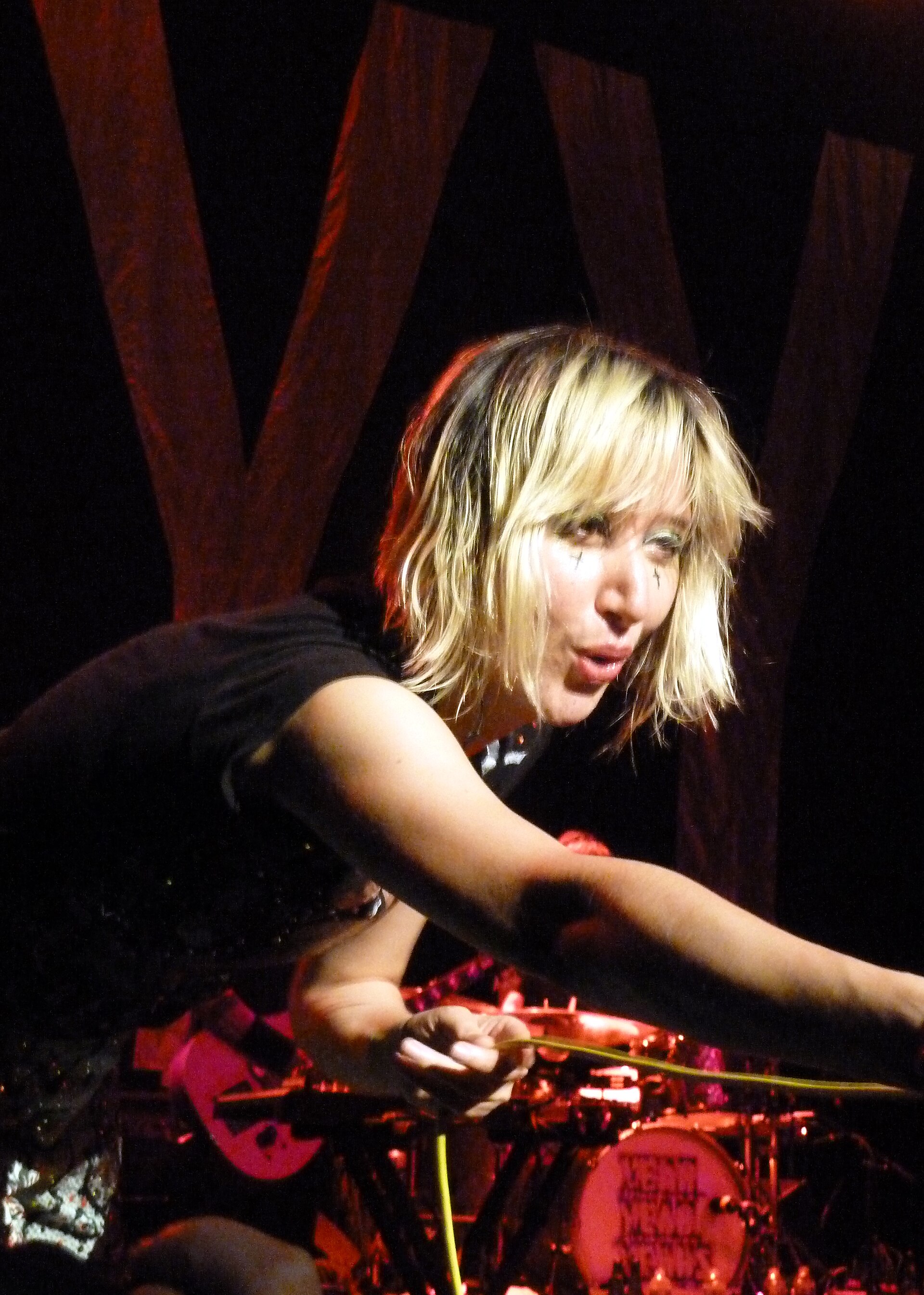 Karen O leans forward over the front of the stage with one arm outstretched, blonde bob falling across painted marks under her eyes.