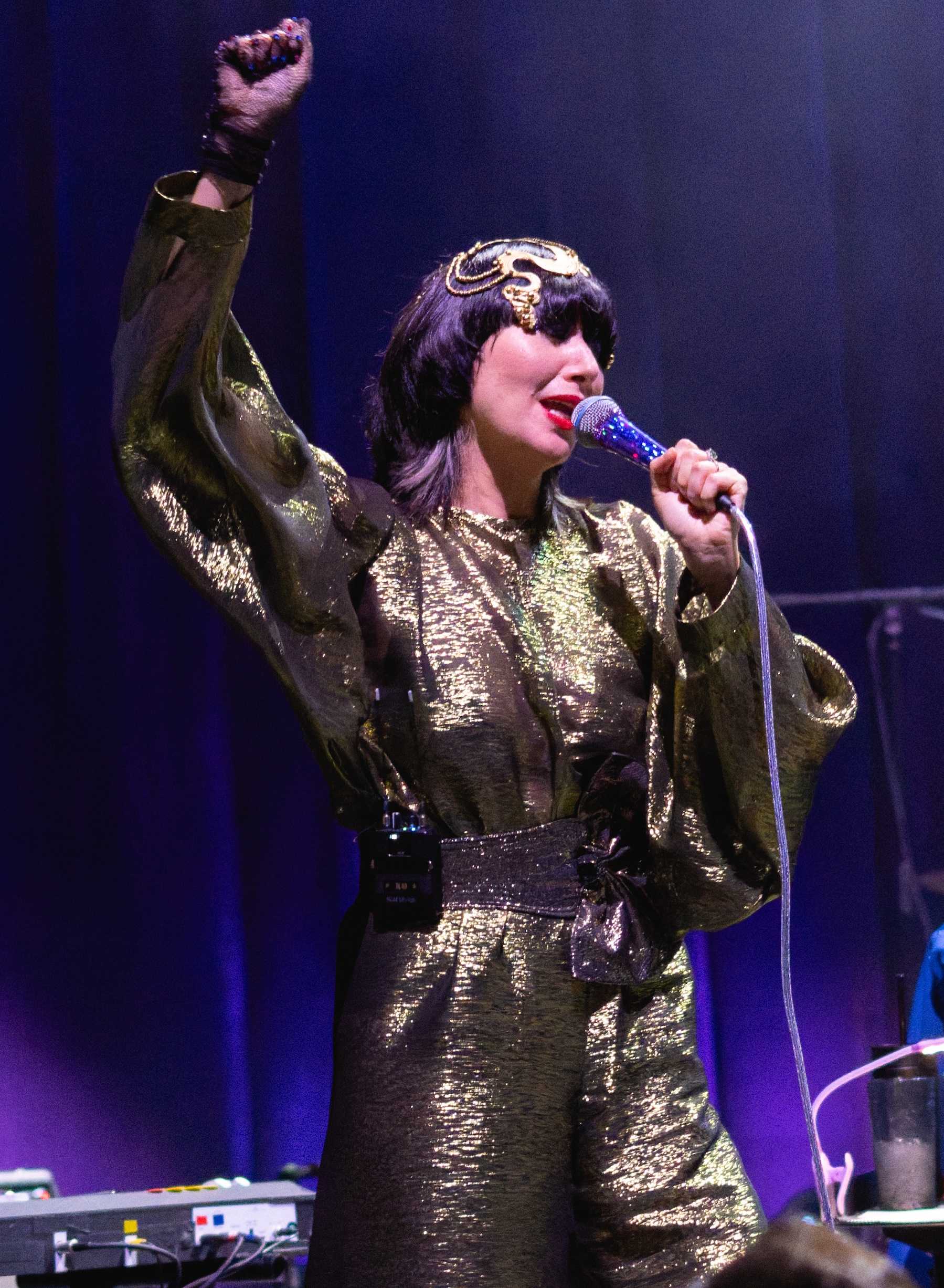 Karen O sings into a corded microphone with one gloved fist raised, wearing a gold lame jumpsuit and a gold headpiece over dark hair.