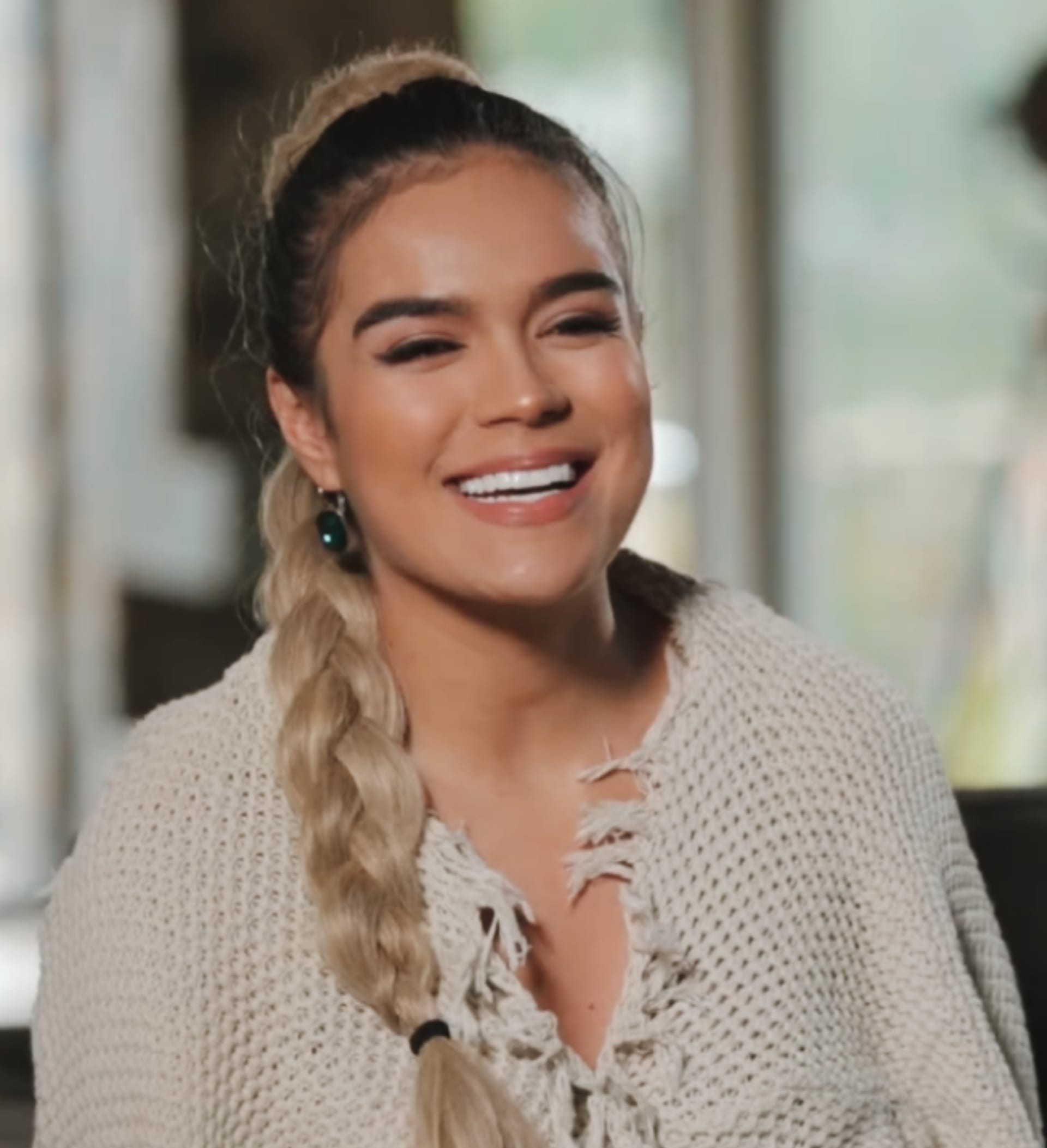 Karol G smiles broadly with her hair in a braided ponytail, wearing a cream knit top during a television interview.