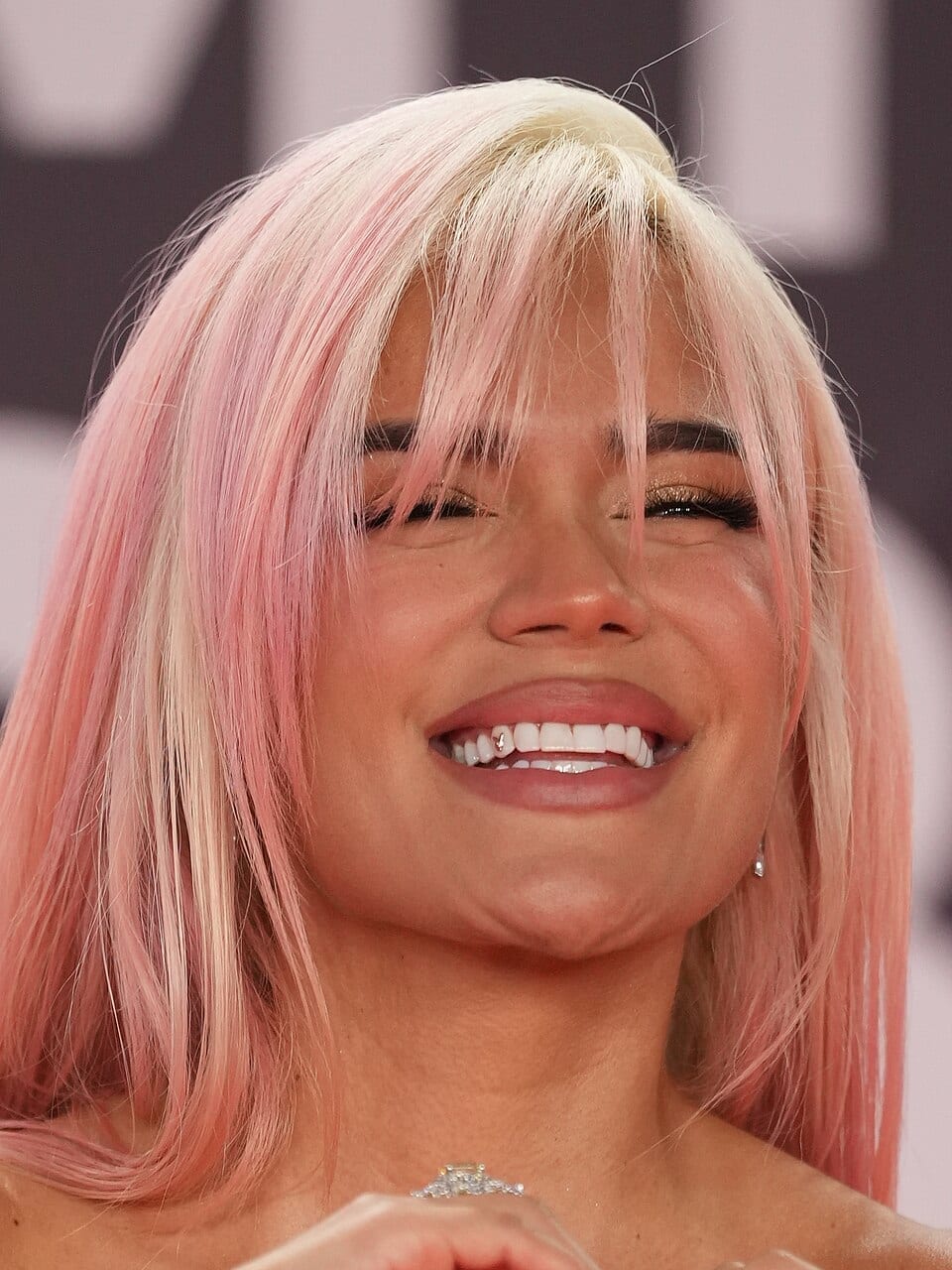 Karol G smiles with her eyes closed, wearing a long pink and blonde wig, at an awards ceremony.