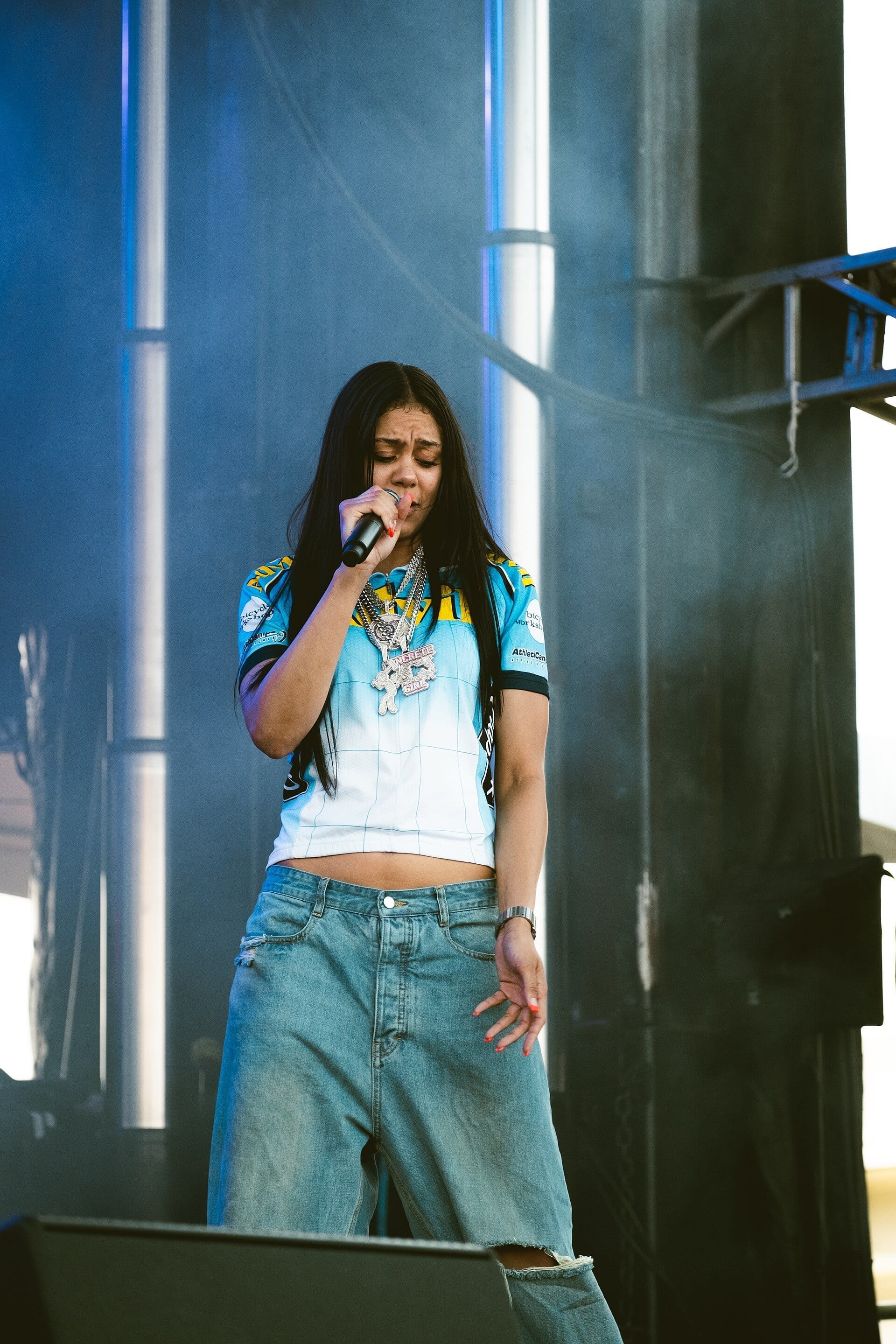 Karrahbooo sings into a microphone on an outdoor festival stage, wearing a sports jersey and layered chain necklaces.