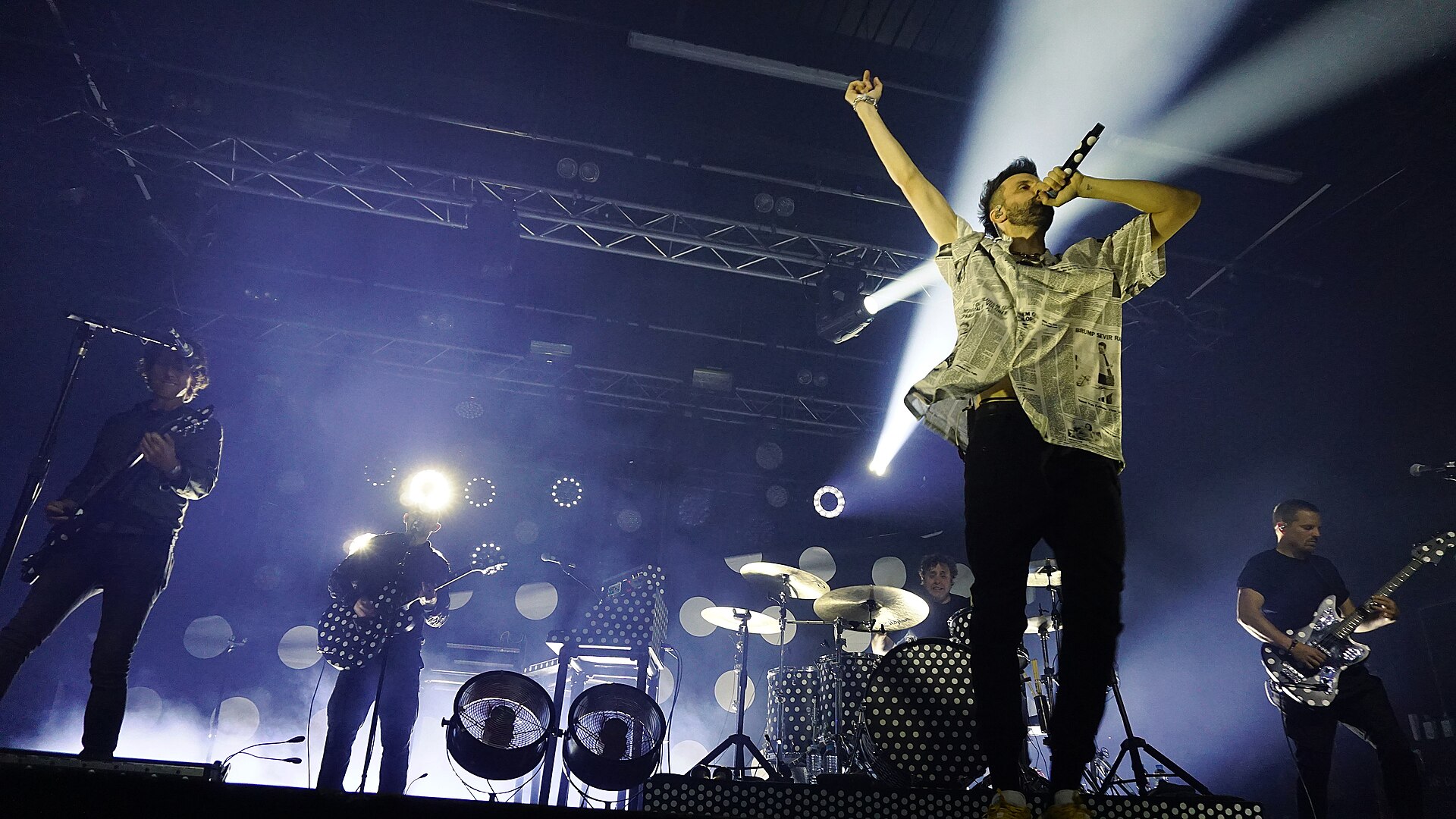 The singer of Kasabian stands on a monitor with both arms thrown up while the rest of the band plays behind him in beams of white light.