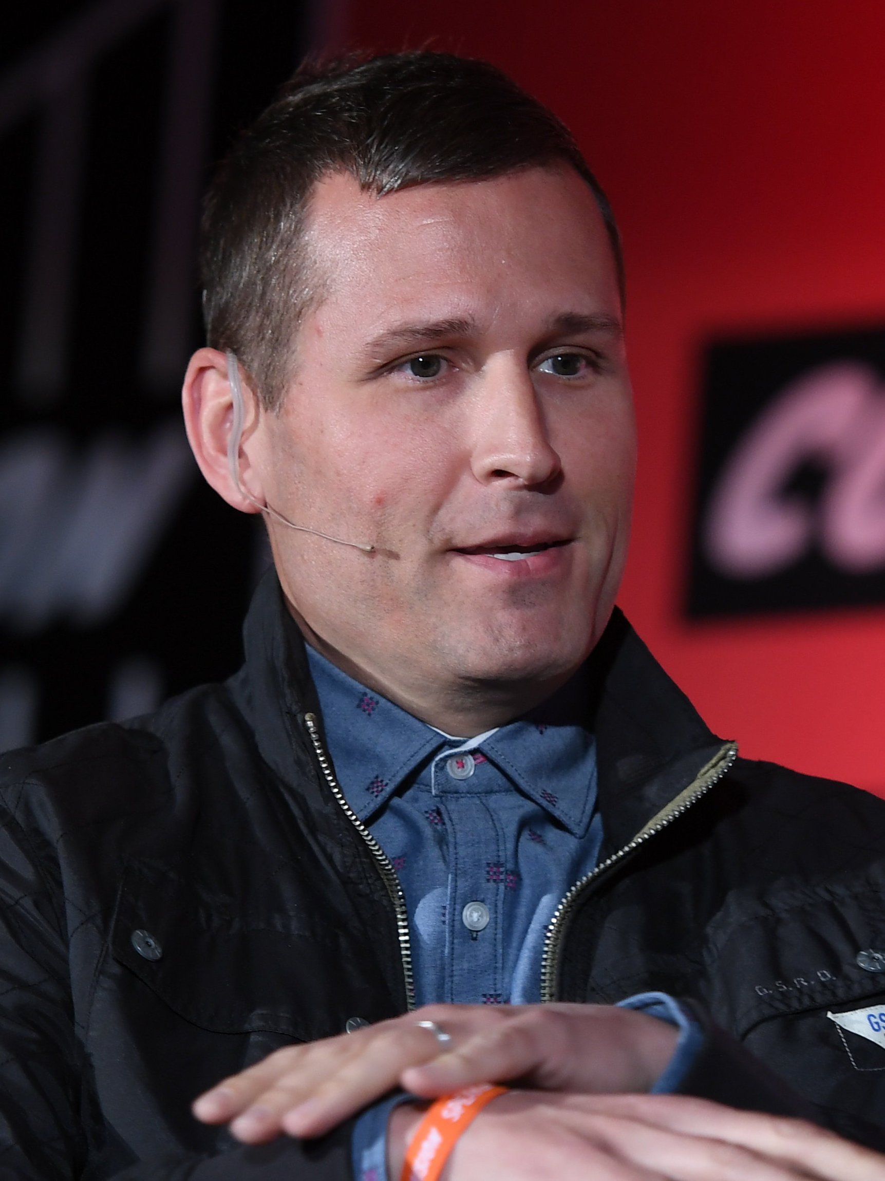 Kaskade speaks into a headset microphone in front of a red backdrop, in a dark button-down shirt.