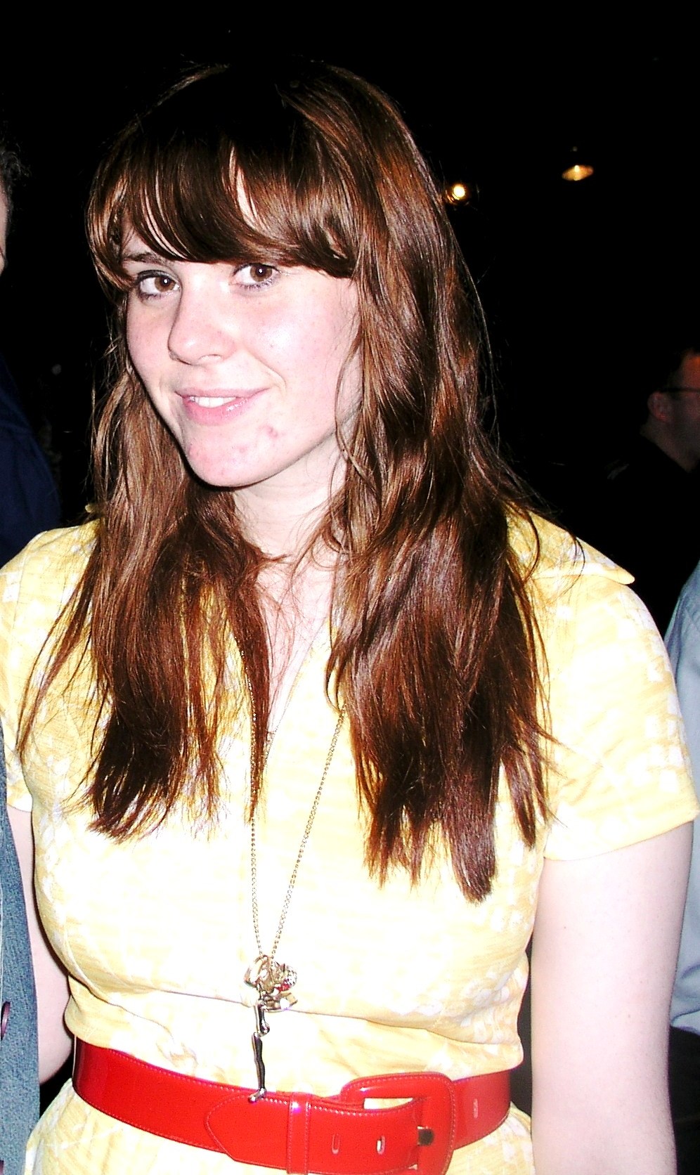 Kate Nash looks at the camera with a half smile, long auburn hair and a heavy fringe, wearing a pale yellow dress, a red patent belt and a long pendant necklace.
