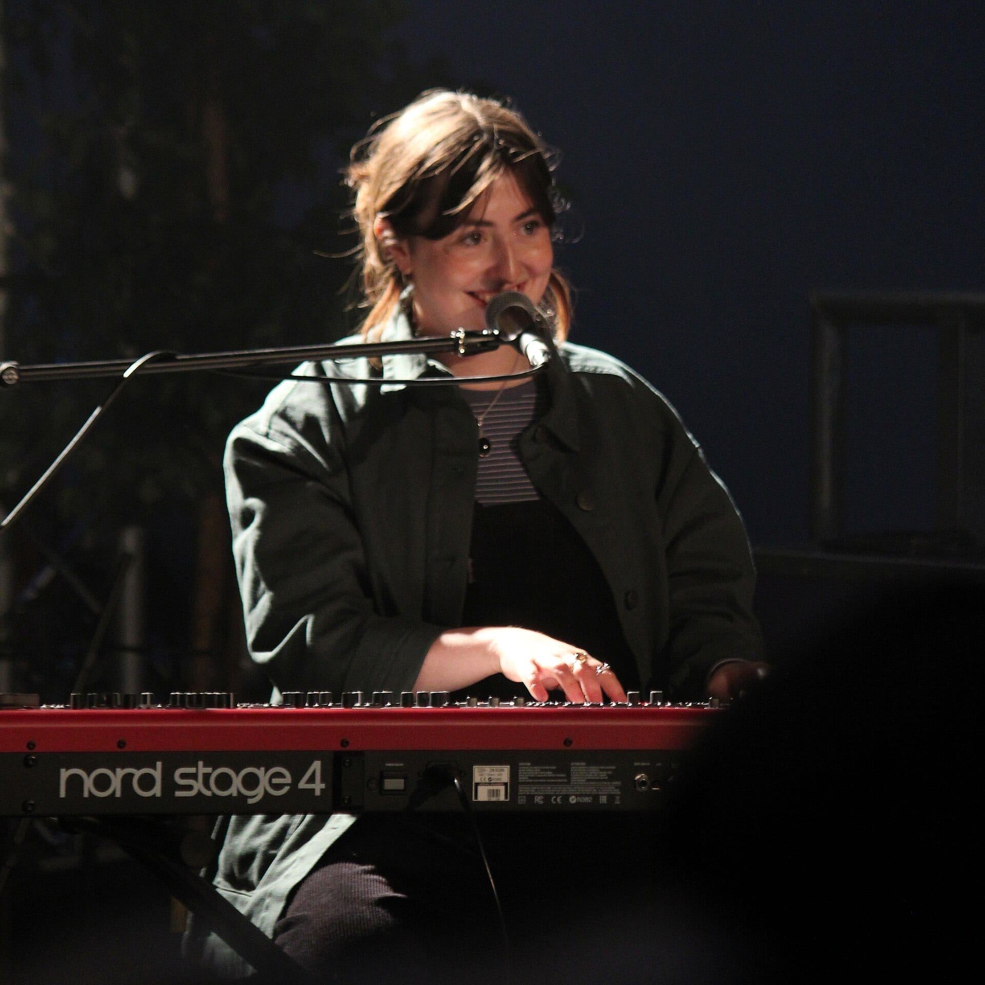 Katie Gregson-Macleod smiles while playing a Nord Stage 4 keyboard behind a microphone stand on a dark stage.