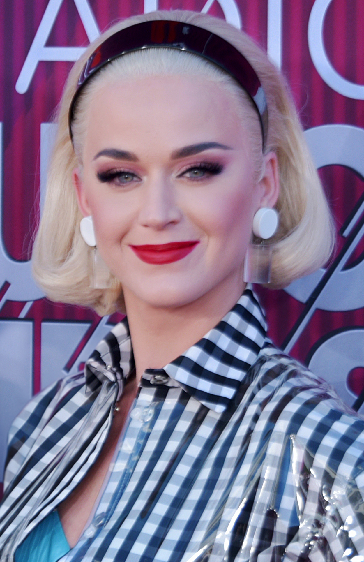 Katy Perry smiles at the camera in a platinum blonde bob and a dark headband, wearing a checked shirt under a clear plastic coat.