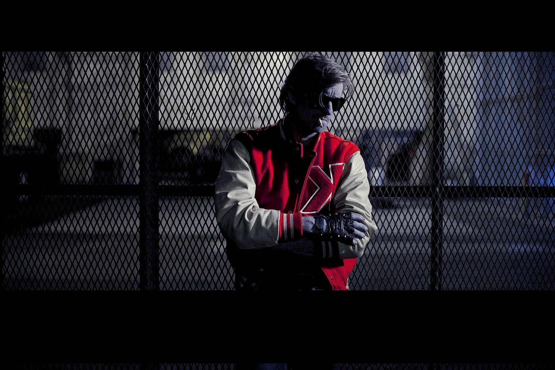 Kavinsky, in sunglasses and his signature red and white varsity jacket, stands with arms crossed behind a chain-link fence at night.