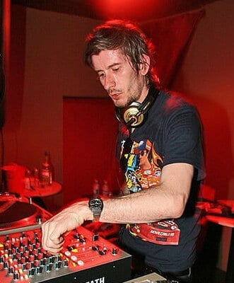 Kavinsky, wearing headphones around his neck, works a DJ mixer under red club lighting.