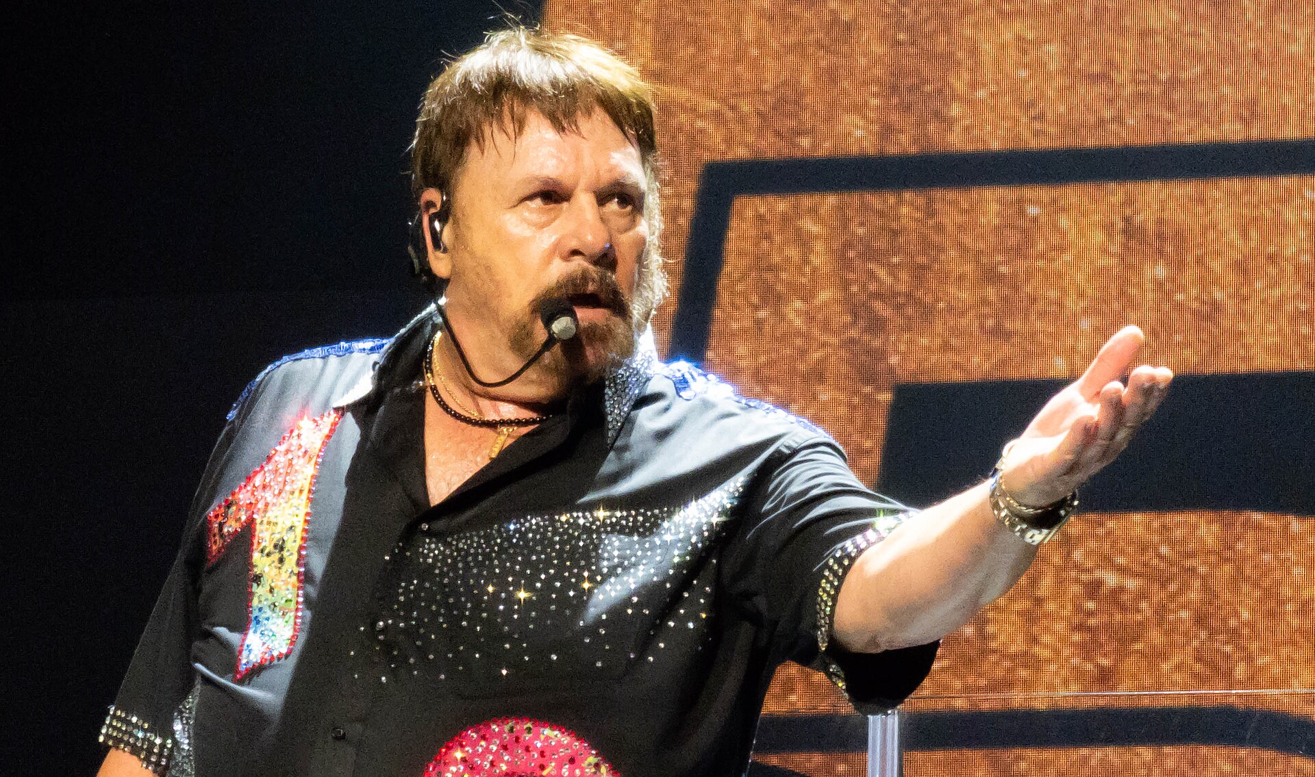 Harry Wayne Casey sings into a headset microphone in a black shirt covered in rhinestones, one arm raised, with a bandmate on guitar behind him.