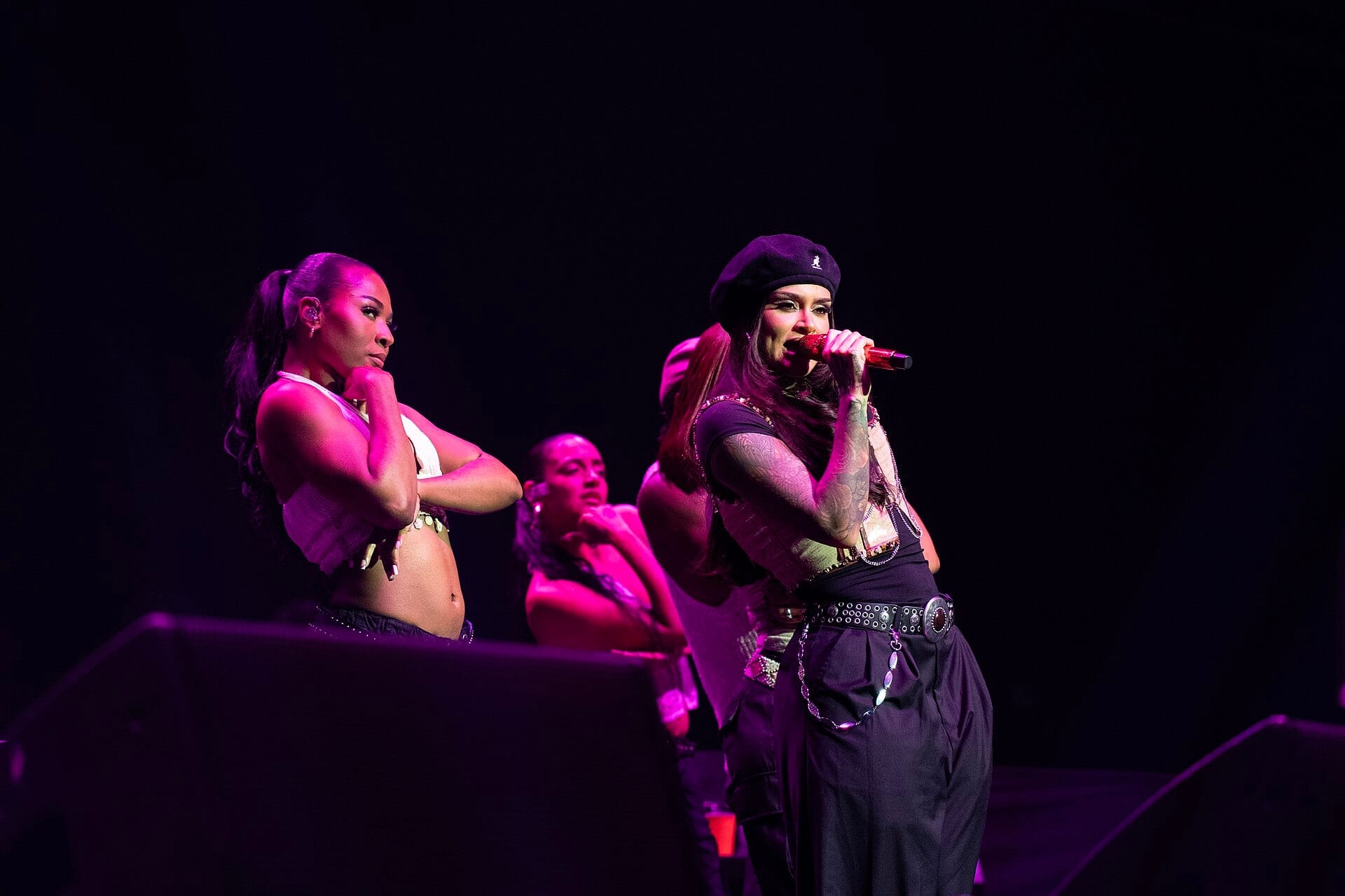 Kehlani sings into a red microphone in a black beret and studded vest, tattooed arms raised, with two dancers posed behind her under magenta light.