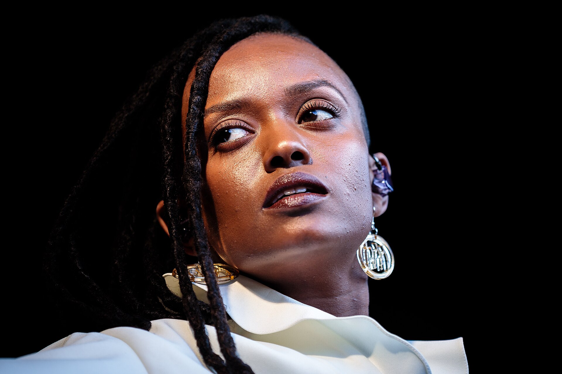 A close-up of Kelela mid-song against a black background, locs falling over one shoulder, large hoop earrings and a white ruffled collar.