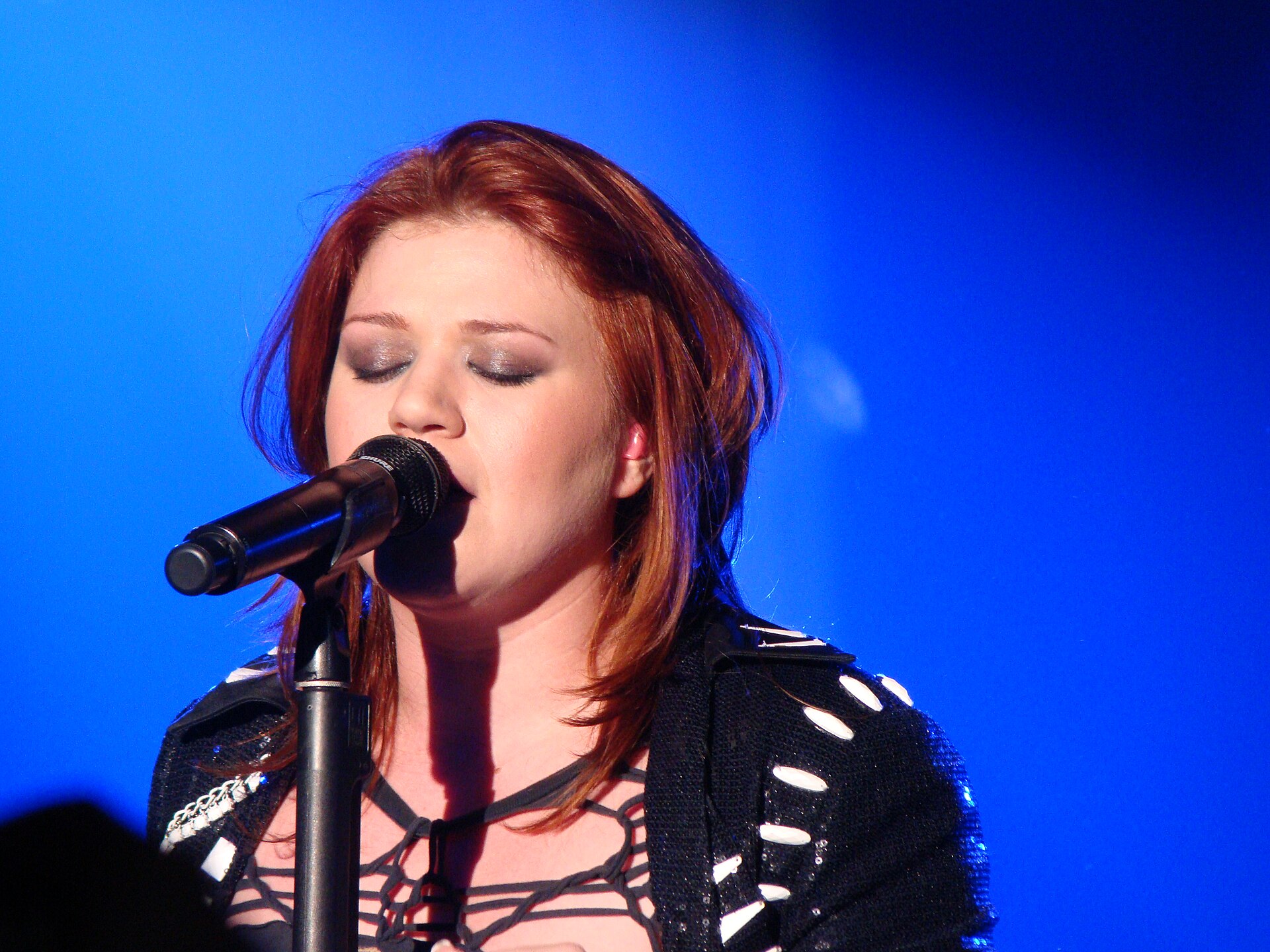 Kelly Clarkson sings into a microphone on a stand with her eyes closed, red hair loose, in a studded black jacket against a deep blue stage backdrop.