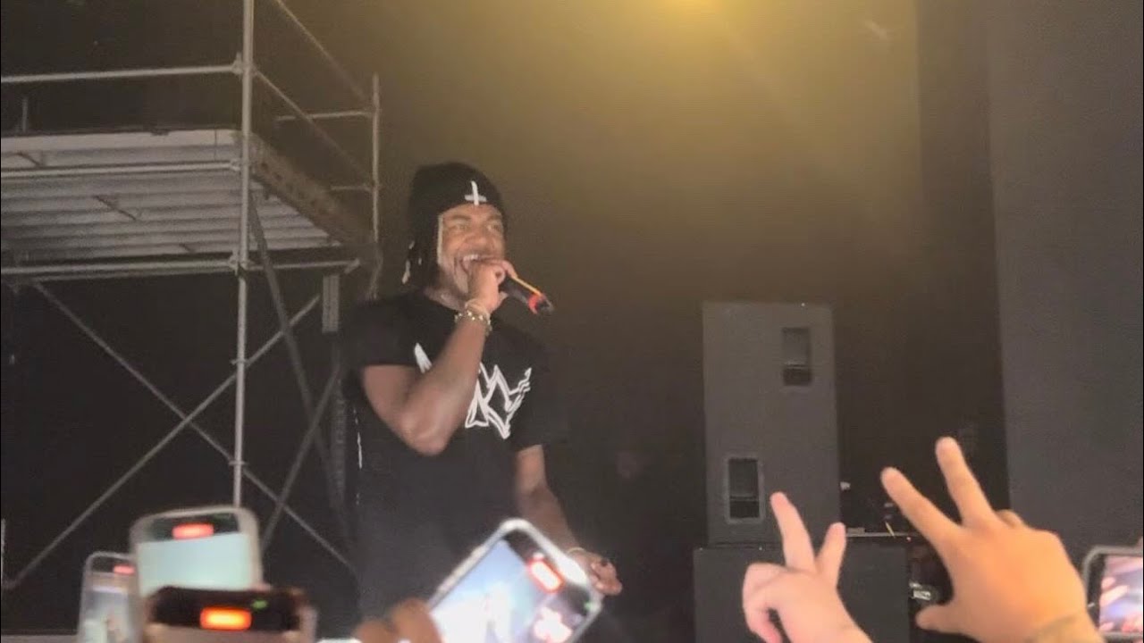 A rapper in a black durag and black T-shirt grins while holding a microphone on a hazy stage, fans' raised phones in the foreground.