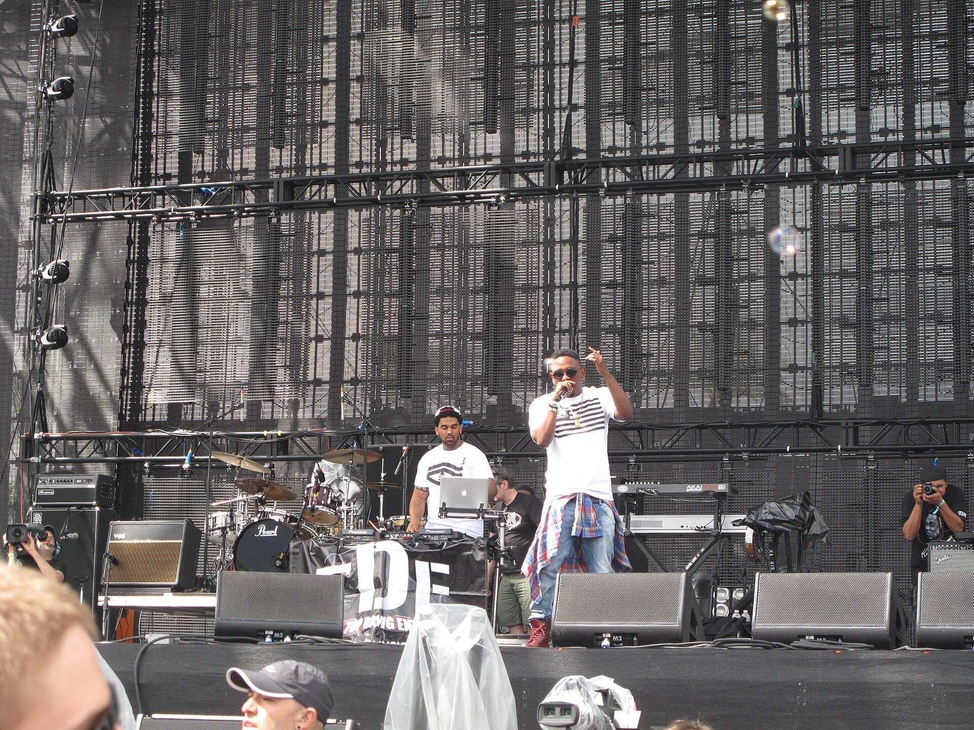 Kendrick Lamar performs on an outdoor festival stage, holding a microphone and wearing sunglasses, a cap, and a striped shirt, with a DJ setup behind him.