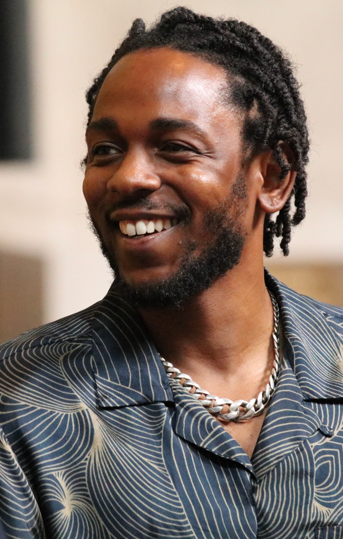 Kendrick Lamar smiles in a patterned short-sleeved shirt with a heavy silver chain, photographed close up indoors.