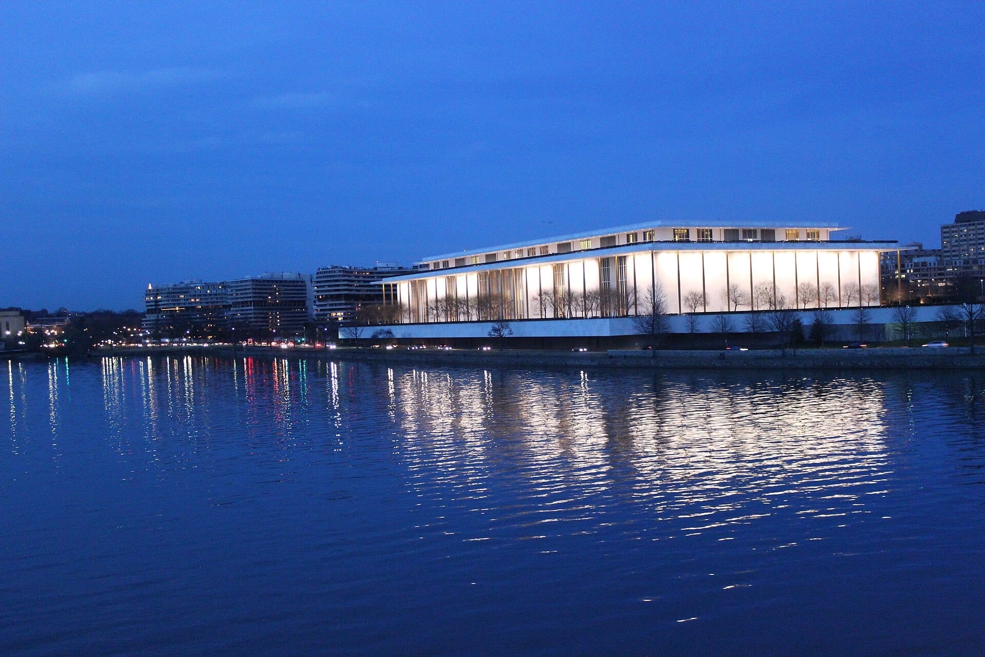The Kennedy Center's illuminated glass facade glows at dusk, reflected in the Potomac River.