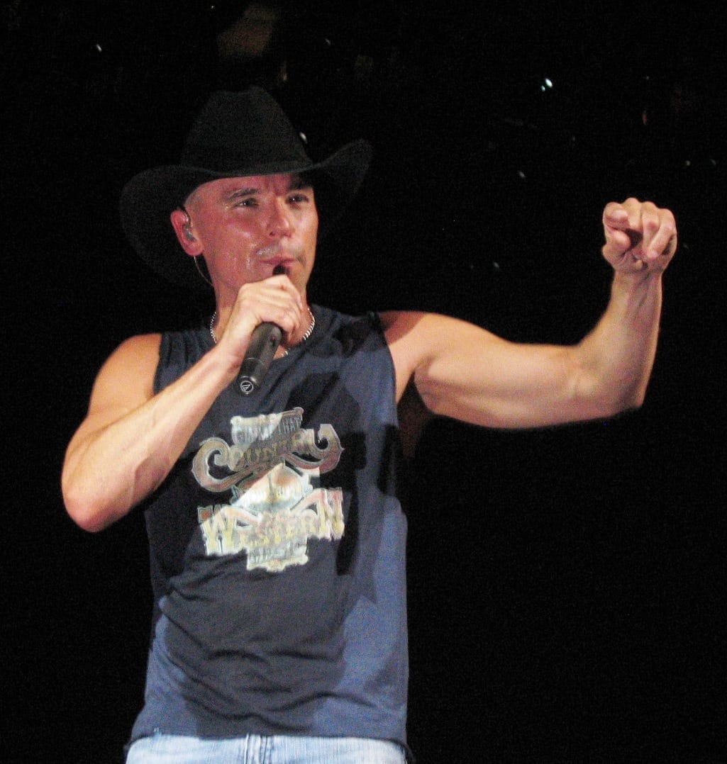 Kenny Chesney sings into a microphone on stage, wearing a black cowboy hat and a sleeveless Country Western tank top.