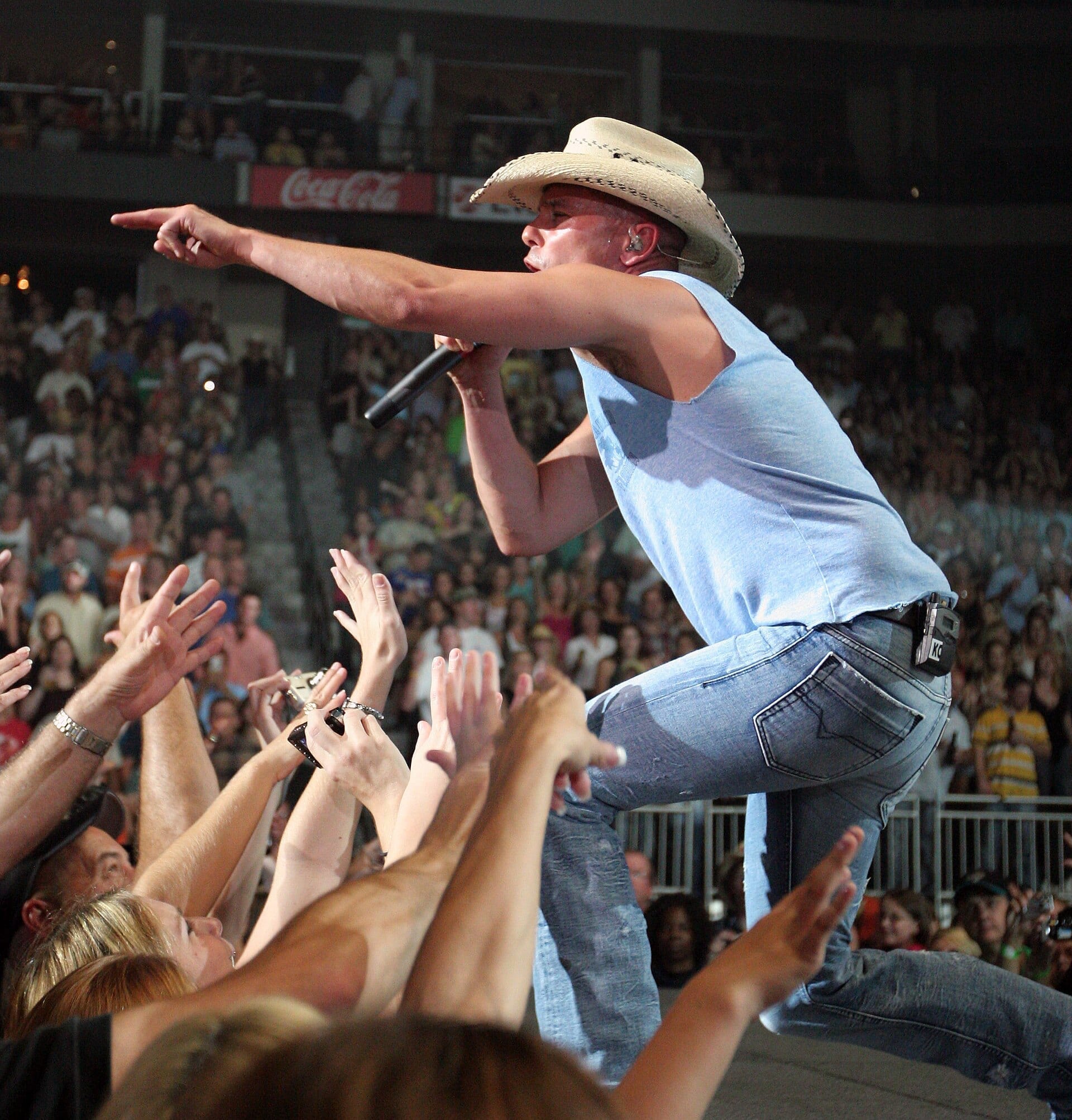 Kenny Chesney leans out over a crowd of reaching hands in a straw cowboy hat and a pale blue tank top, pointing with the microphone hand.