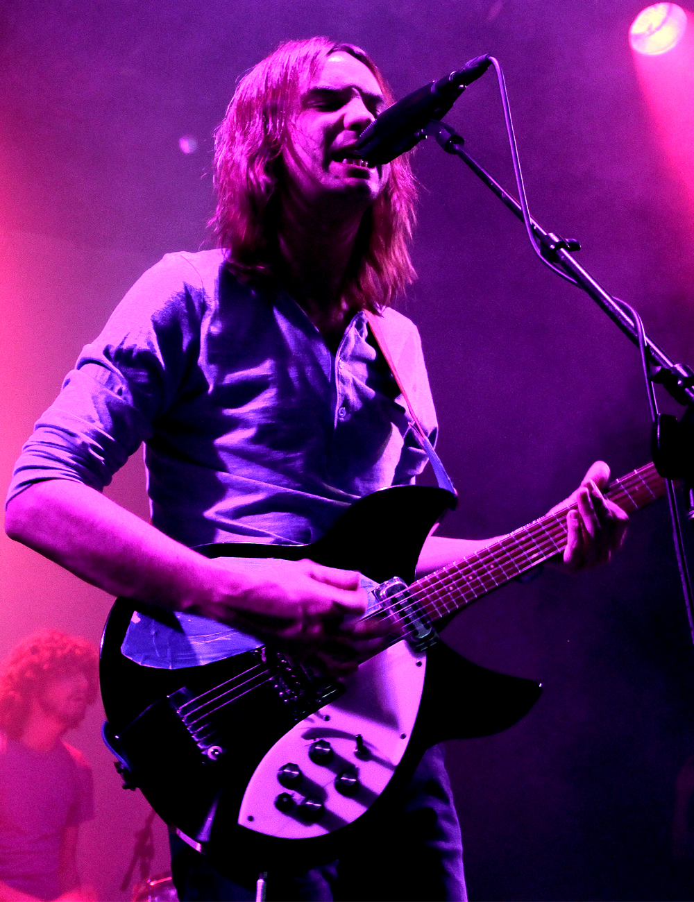 Kevin Parker sings into a microphone while playing a black-and-white Rickenbacker guitar, long fair hair over his face, under magenta stage light.