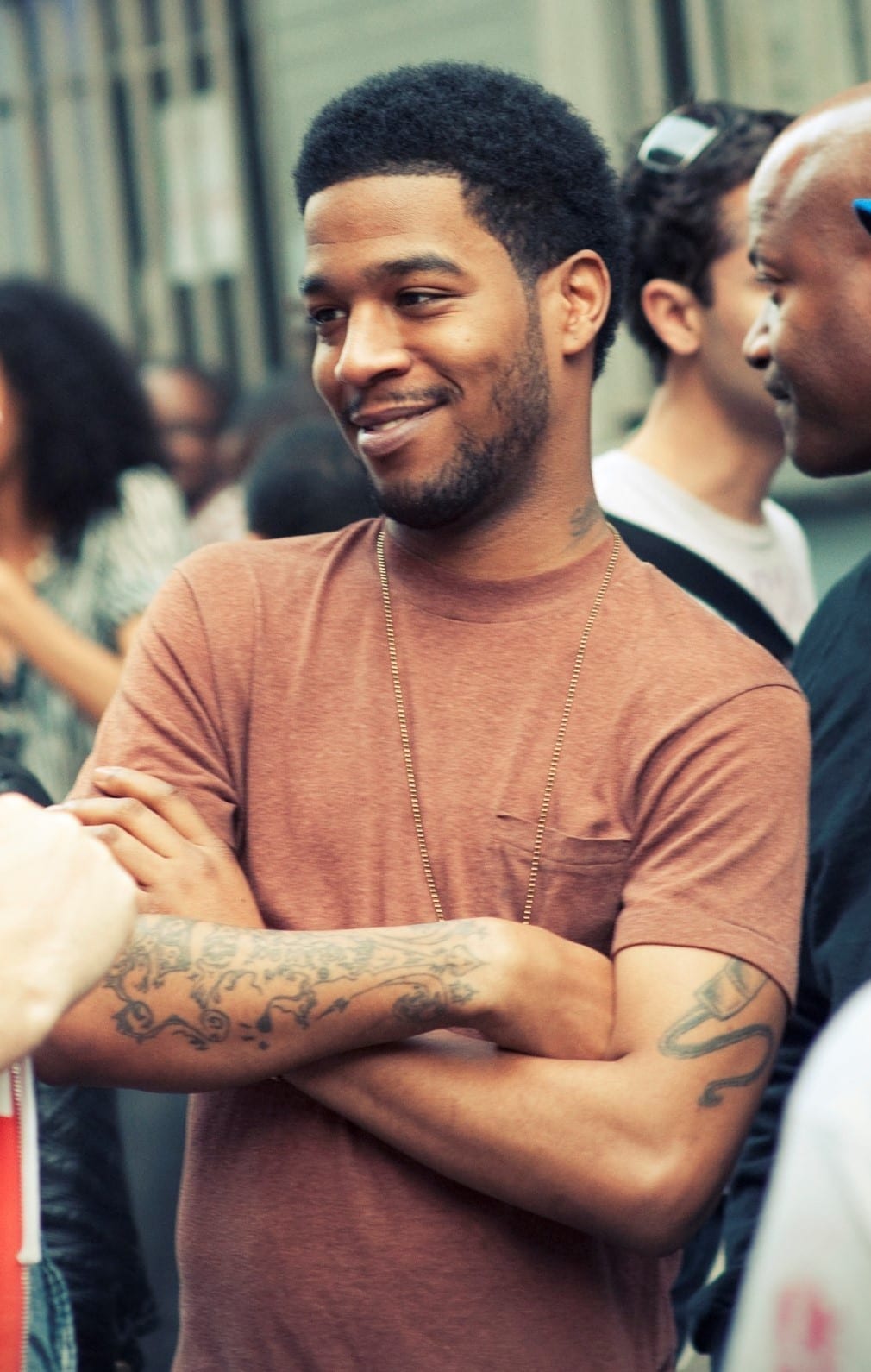 Kid Cudi, arms crossed and smiling, wears a heather rust T-shirt and a long gold chain while standing in a crowd, tattoos visible on both forearms.