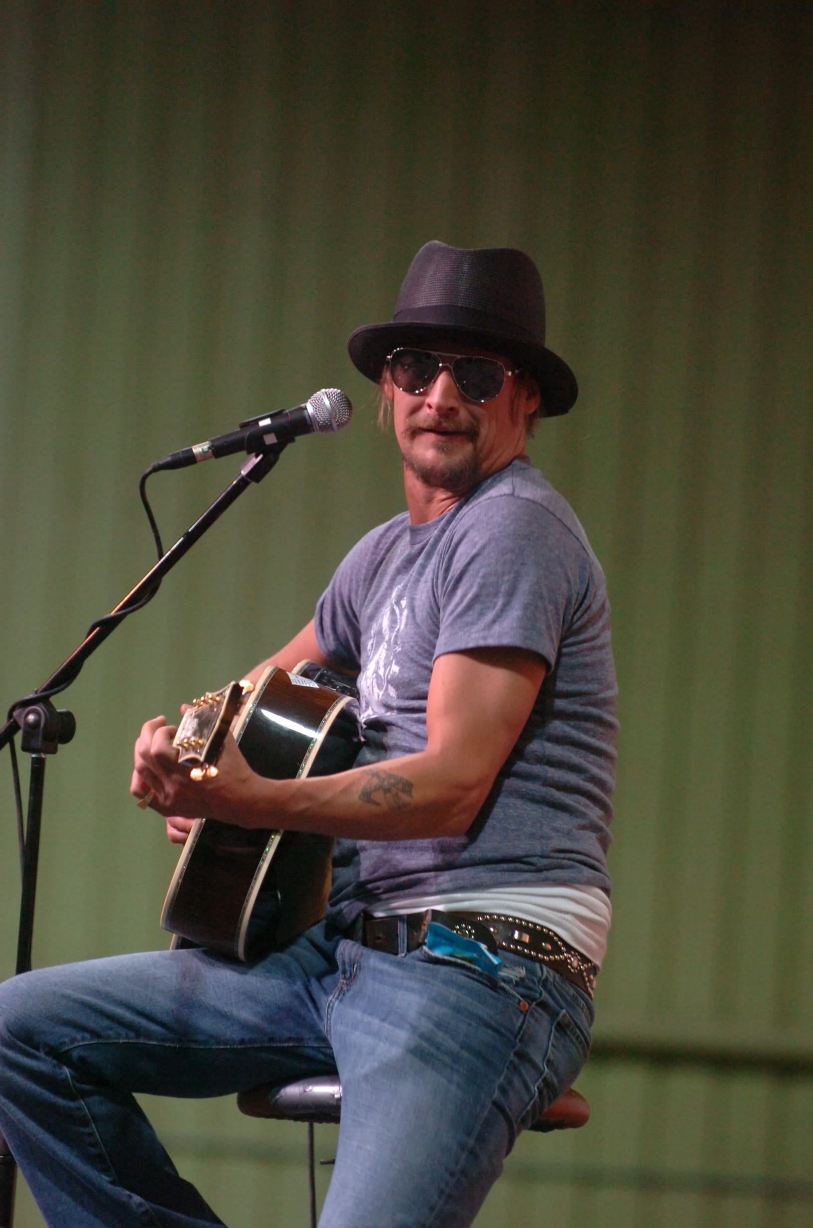 Kid Rock sits on a stool with an acoustic guitar, wearing a black trilby, aviator sunglasses and a grey T-shirt.