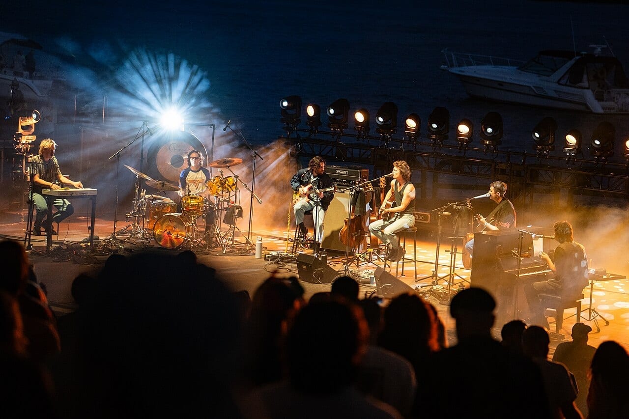 King Gizzard & the Lizard Wizard play an acoustic set on an outdoor stage, with the audience silhouetted in front of them.